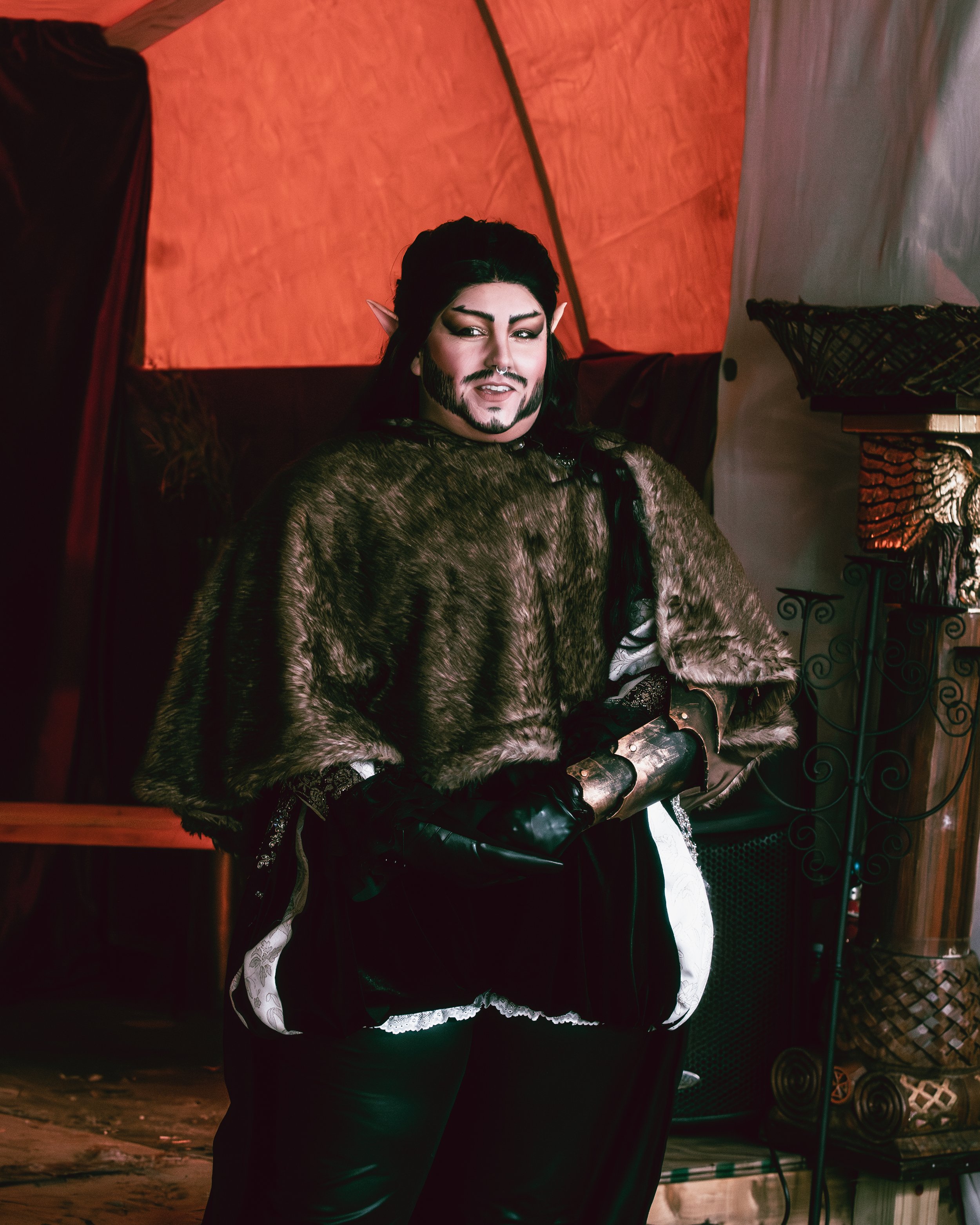 Person dressed as an elf with pointed ears, dark hair, beard, wearing fur cloak, black gloves, and medieval-style outfit, standing in a fantasy-themed setting with red lighting and decorative elements.