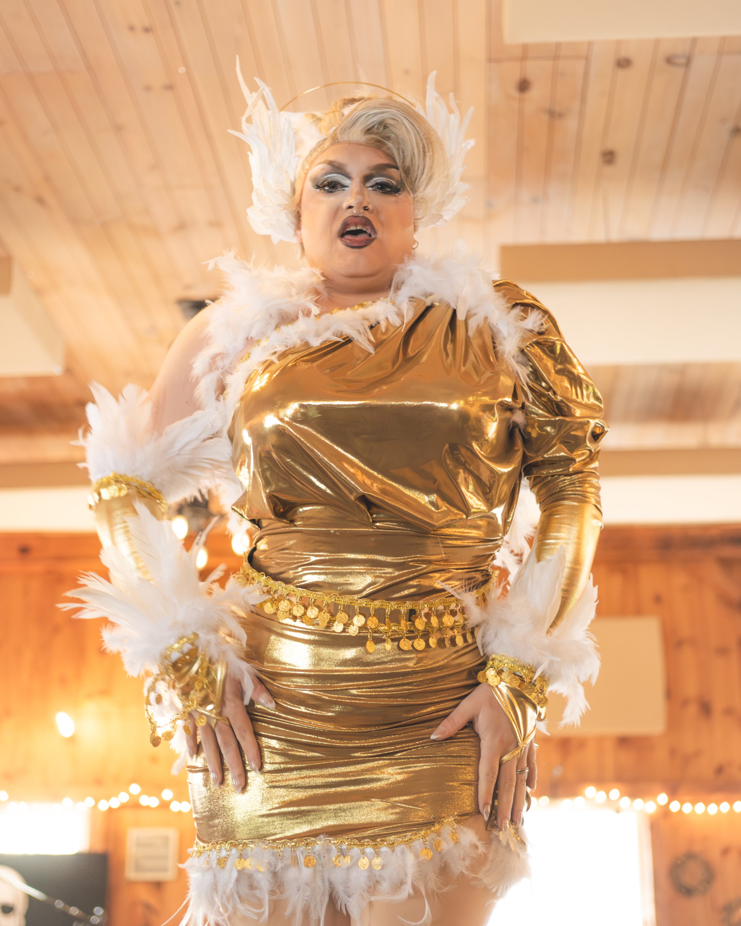Performer dressed in a shiny gold outfit with feathers and jewelry, standing indoors with a wooden ceiling and string lights in the background.