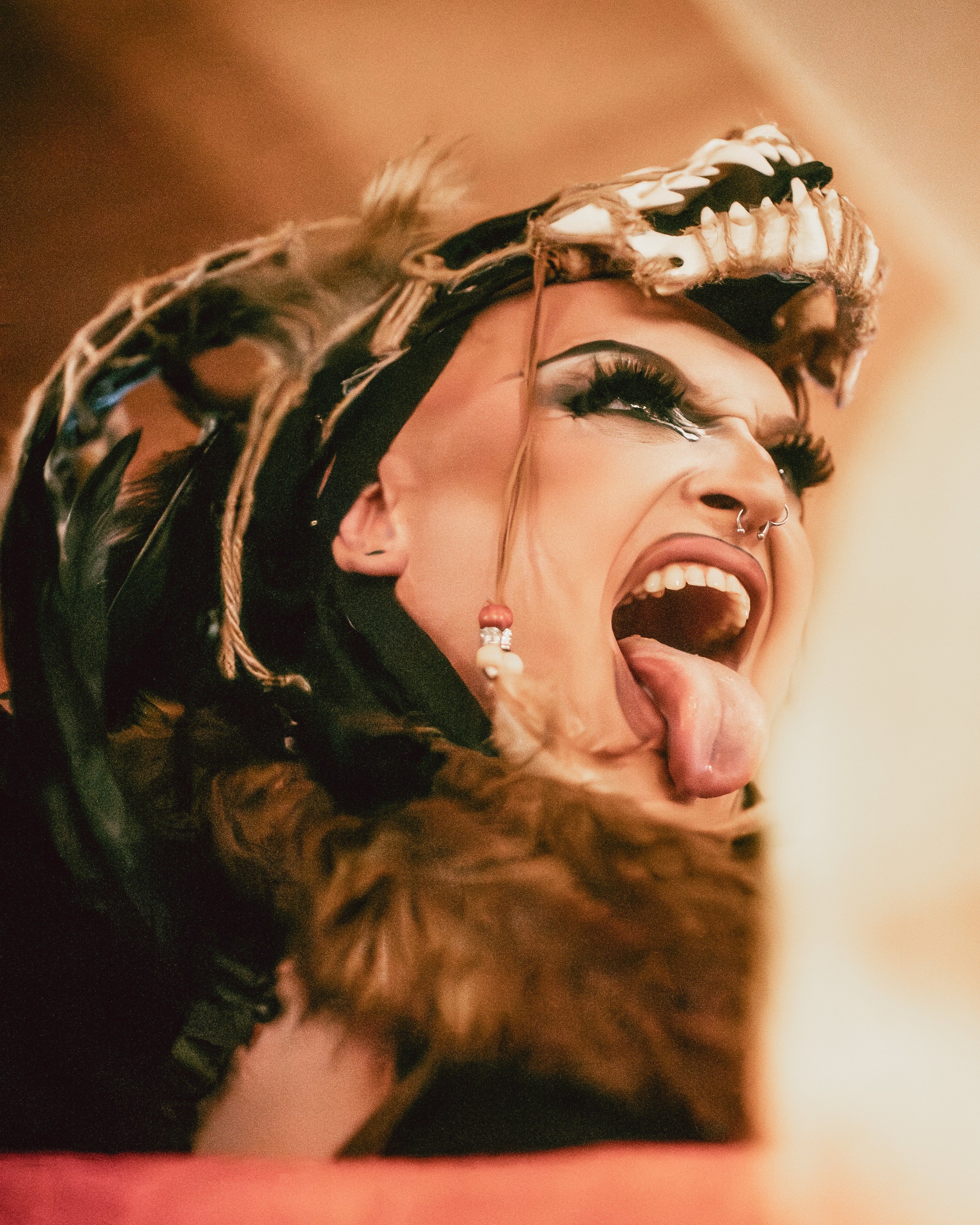 A person wearing a headdress made of feathers and animal skulls with their mouth wide open and tongue sticking out.