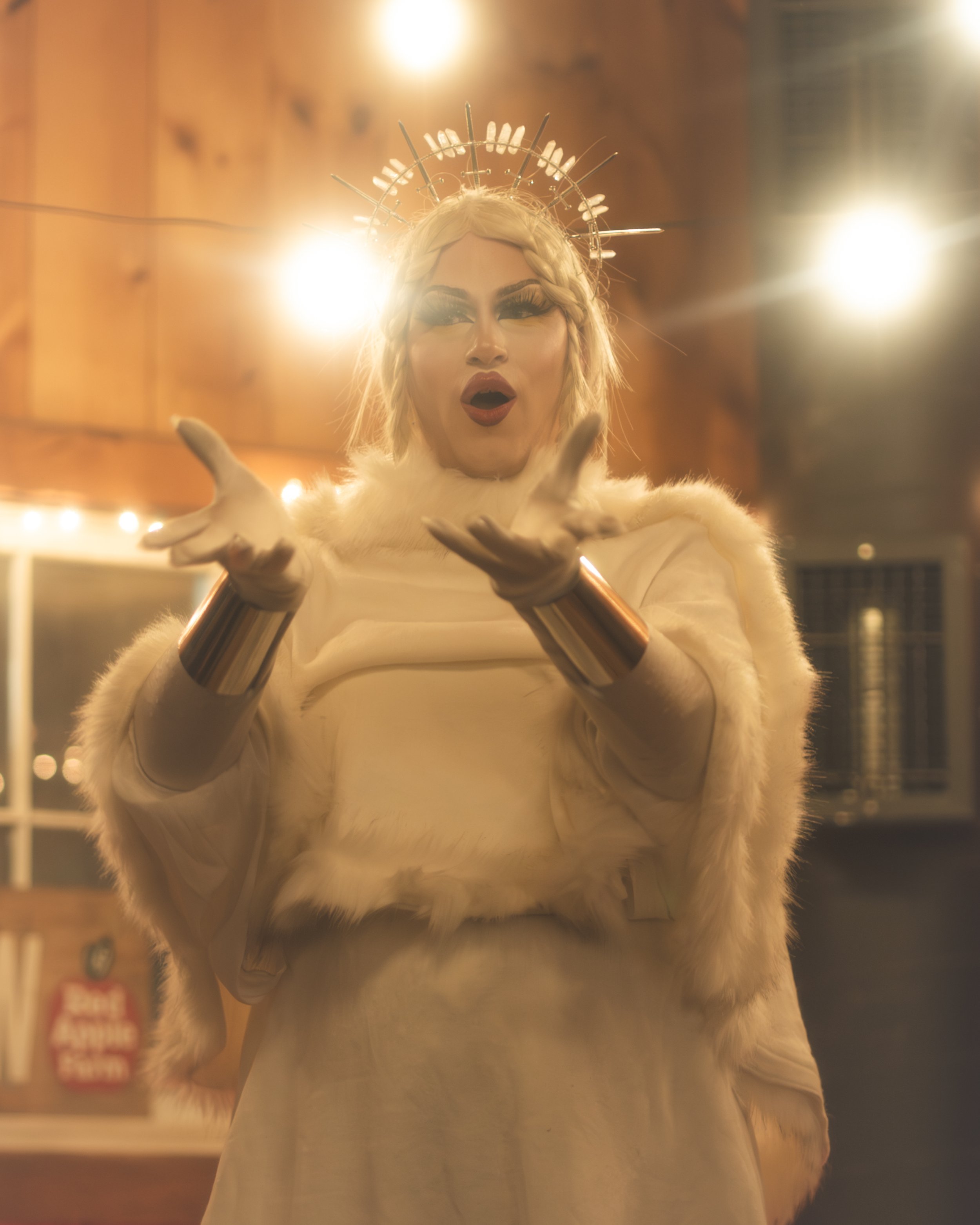 A person dressed in a white, fur-trimmed outfit wearing a headpiece with nails, with arms extended and hands open, in front of a warm, wooden background with lights.