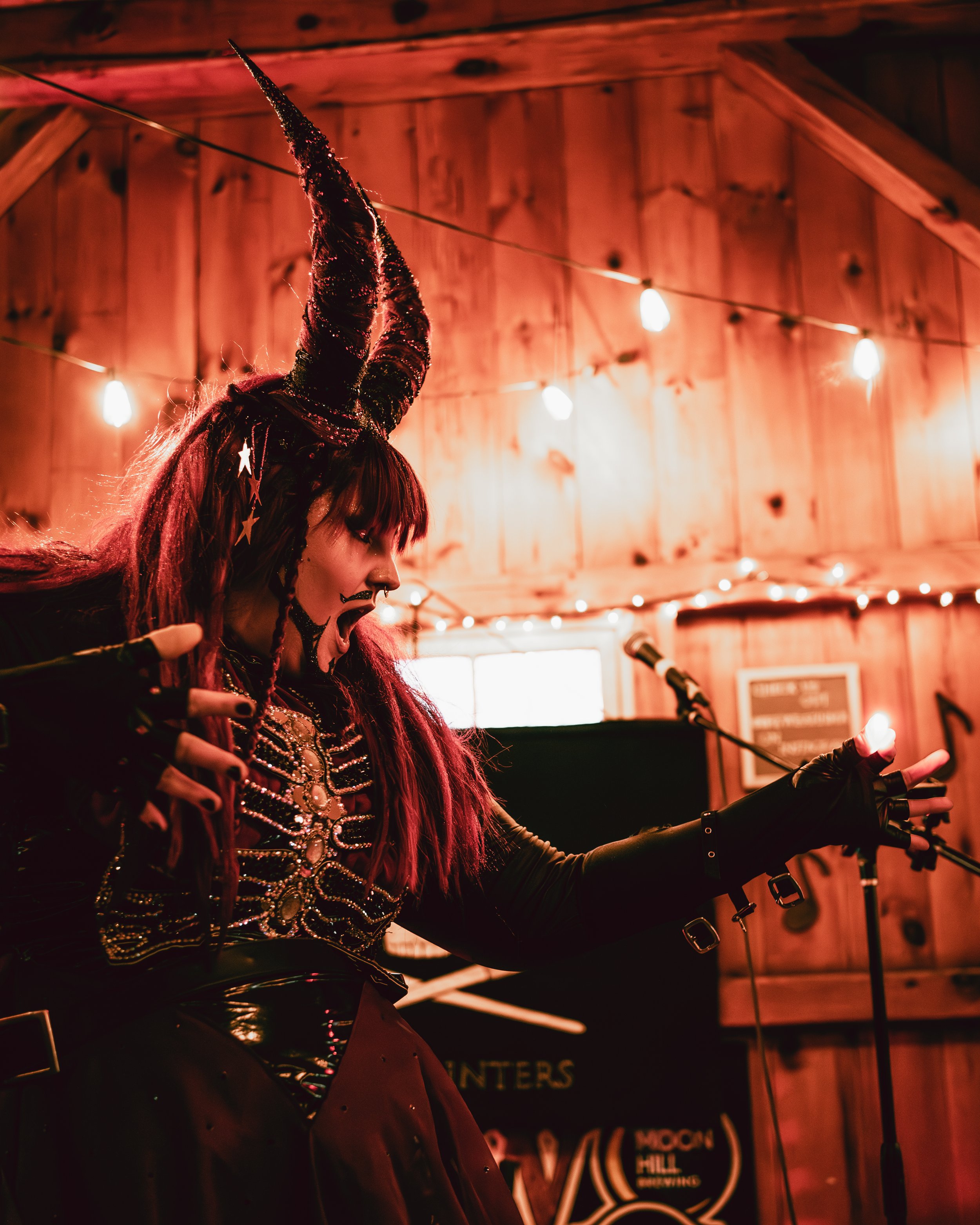 A woman dressed as a witch or fantasy character with a tall, glittery horned hat, face makeup, and long red hair, speaking or singing into a microphone on a wooden stage decorated with string lights.