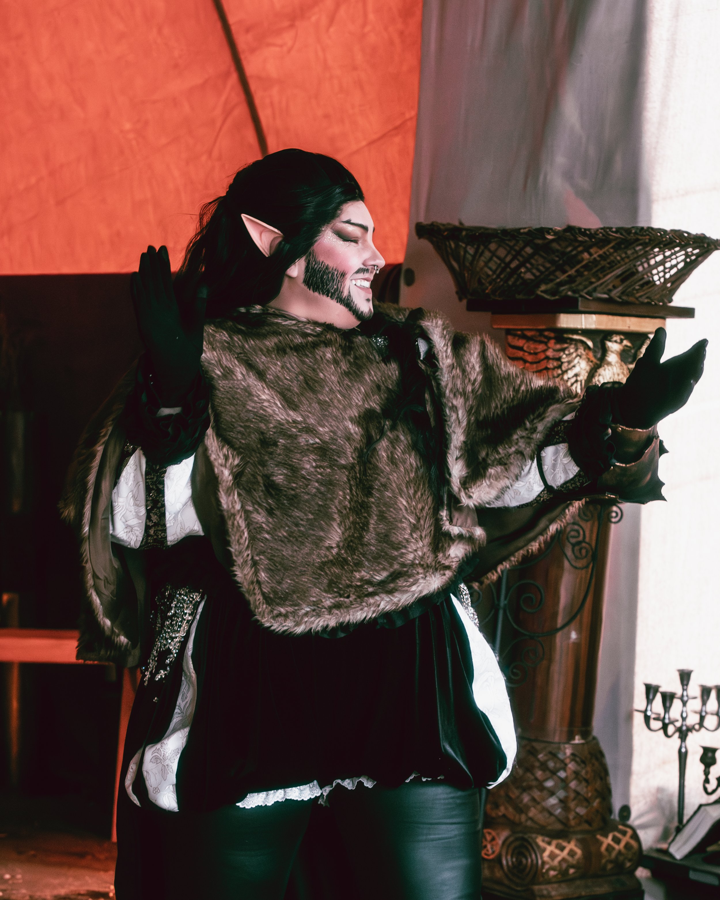 Person dressed as a fantasy elf, smiling and gesturing, wearing a fur cloak, black gloves, and leather pants, with pointed elf ears and dark hair.