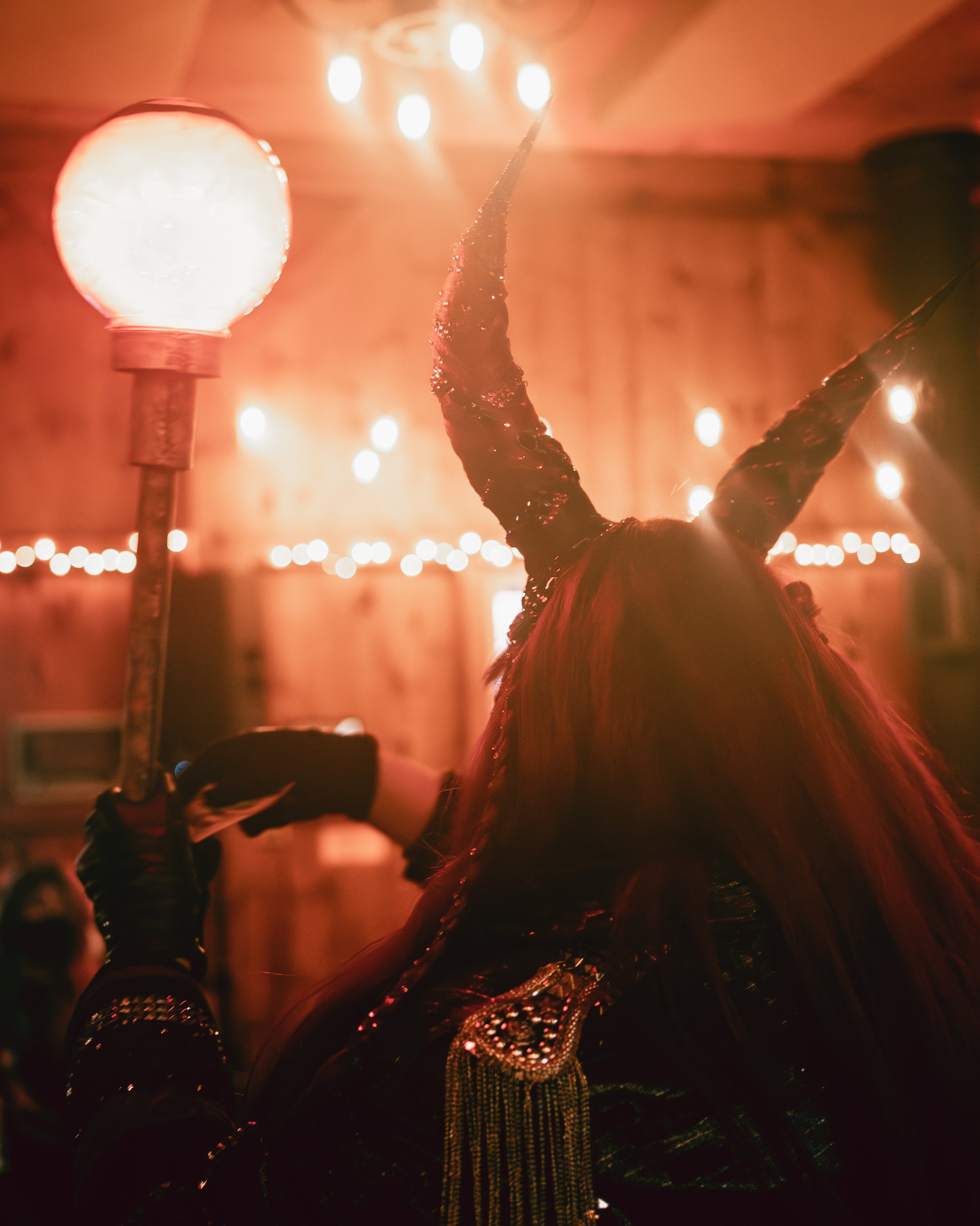 Person dressed as Maleficent holding a staff with a glowing orb, in a warmly lit indoor setting with string lights.
