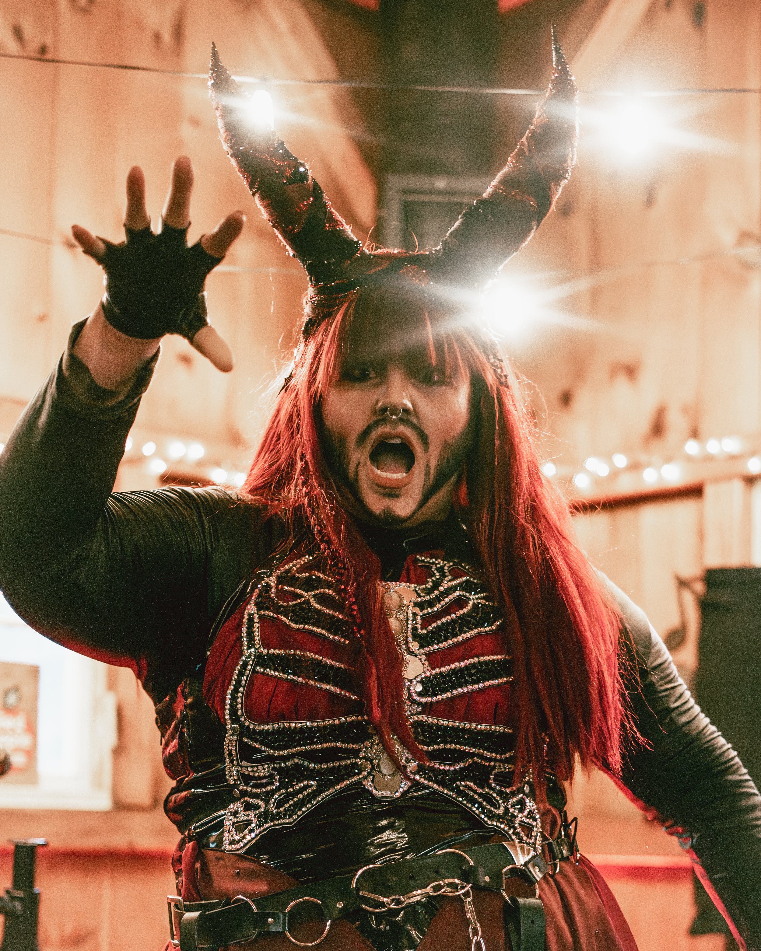 Person with red hair, wearing black and red outfit with silver embellishments, black gloves, and black horned headpiece, making a fierce gesture inside a wooden-paneled room.