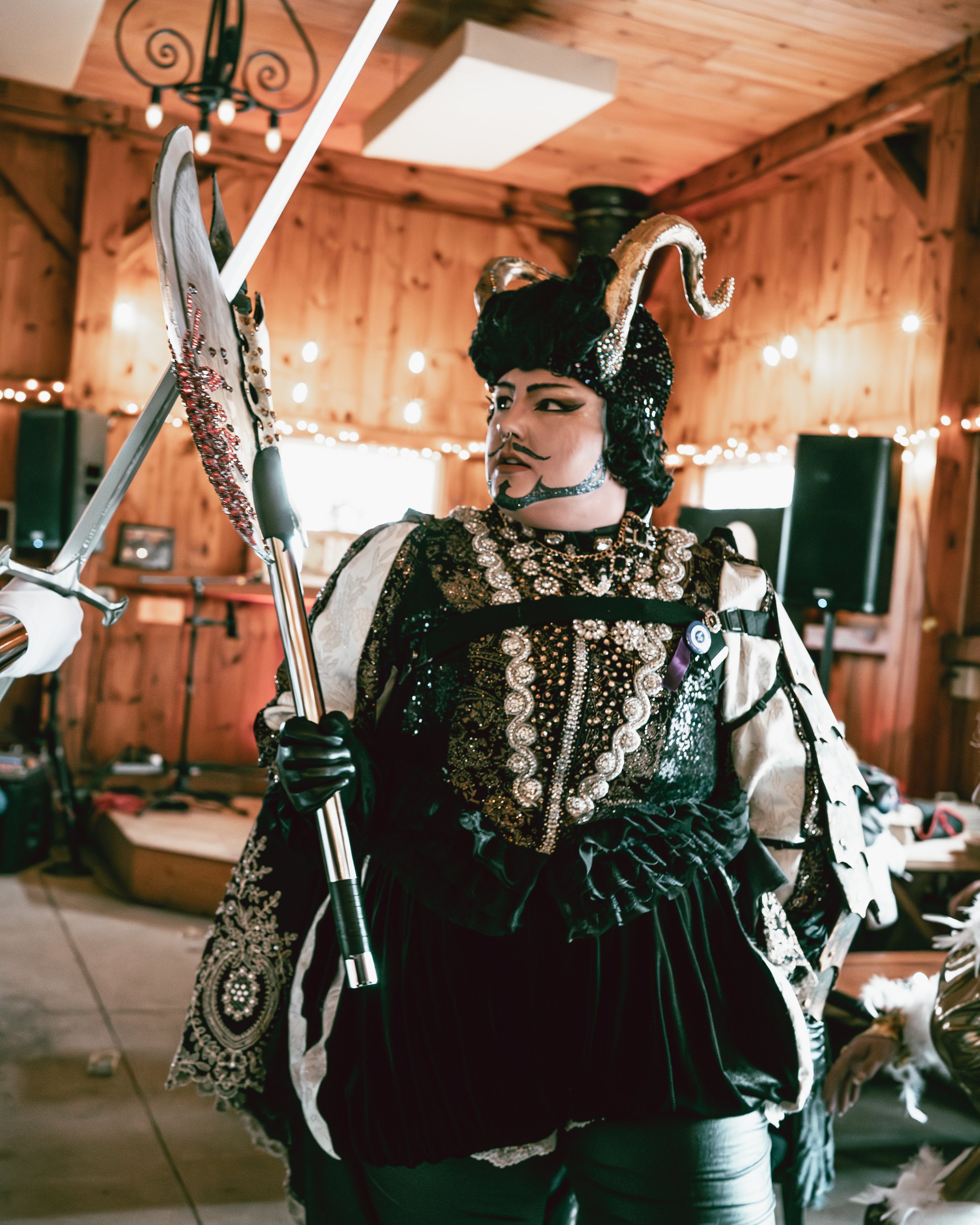 Person dressed in an elaborate, dark-colored costume with intricate embroidery, holding a lance, and wearing a headpiece with large, curled horns and decorative details, indoors with wooden walls and string lights.