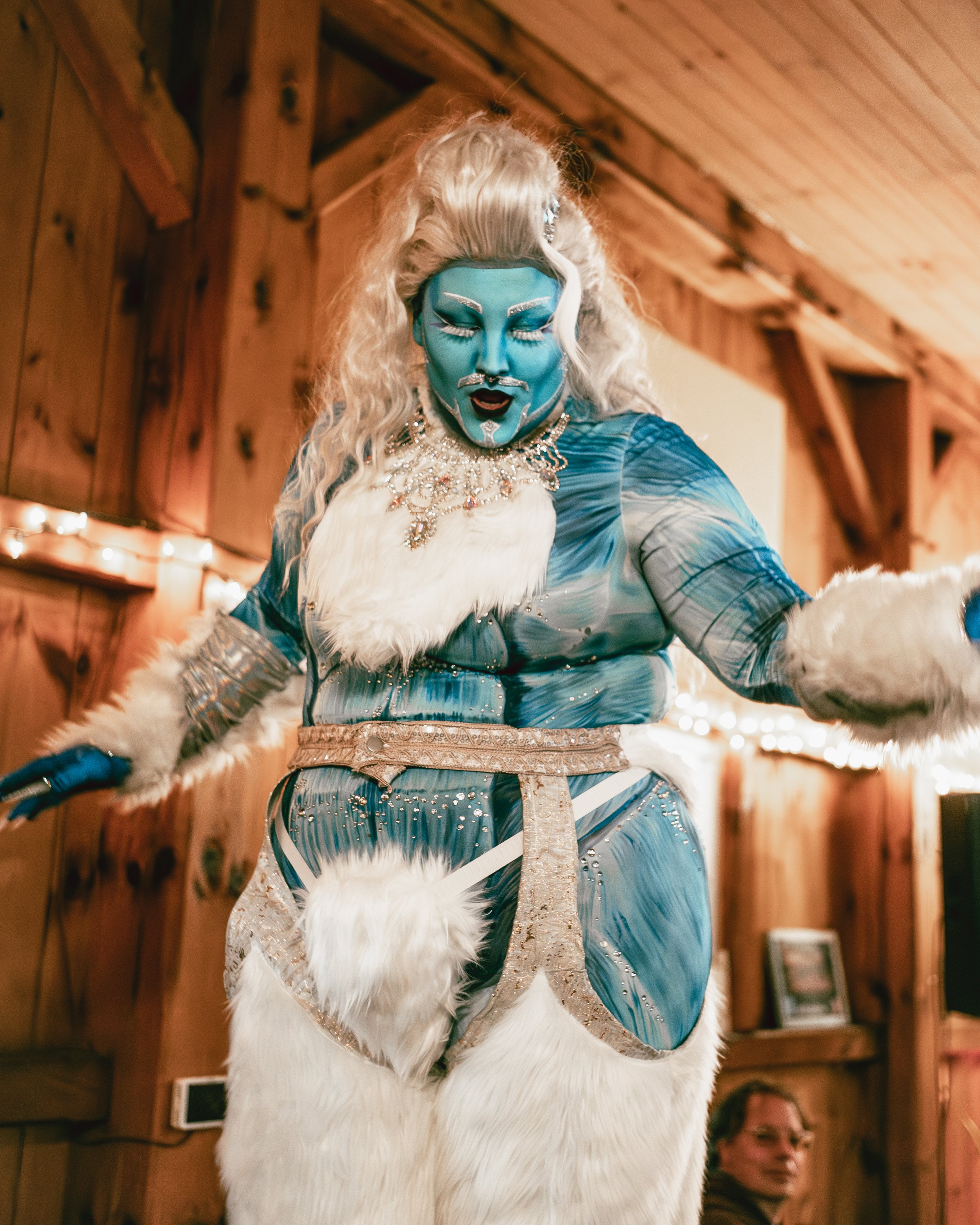 Person in elaborate blue and white costume with face paint and jewelry, performing in a wooden indoor setting.
