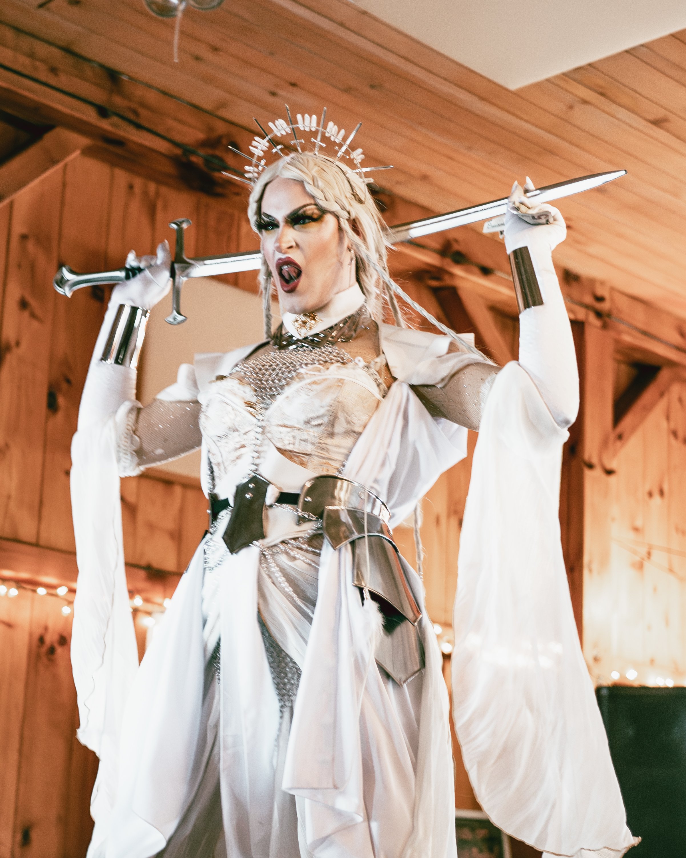 Person dressed as a warrior princess holding a sword over her shoulders, wearing a metallic crown, with a wooden interior background.