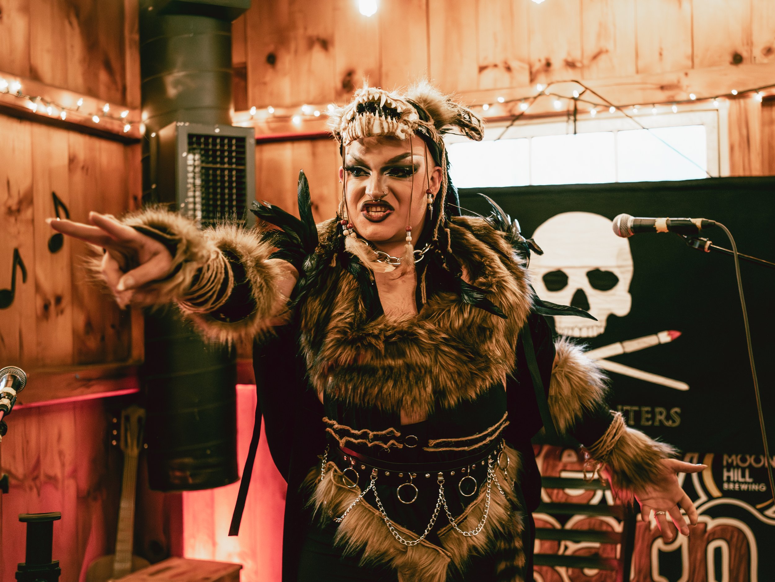 Person dressed in a fierce, animal-inspired costume with fur, chains, and feathers, performing passionately in front of a skull banner at a themed event or performance.
