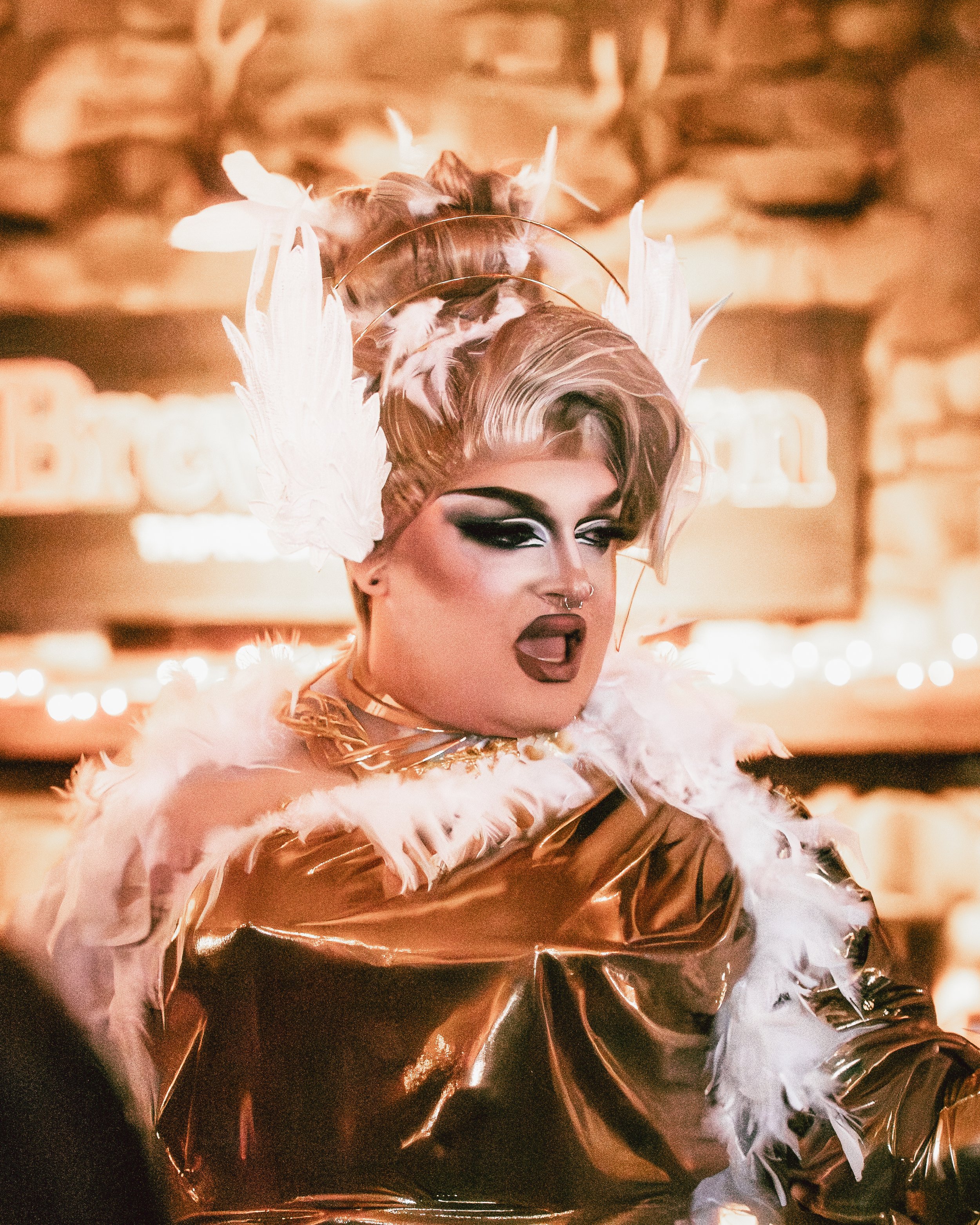 A drag performer with dramatic makeup, a septum piercing, and styled hair in an elaborate feathered headdress, wearing a shiny metallic outfit and feathered accessories, performing or posing in an indoor setting with blurred background lights.