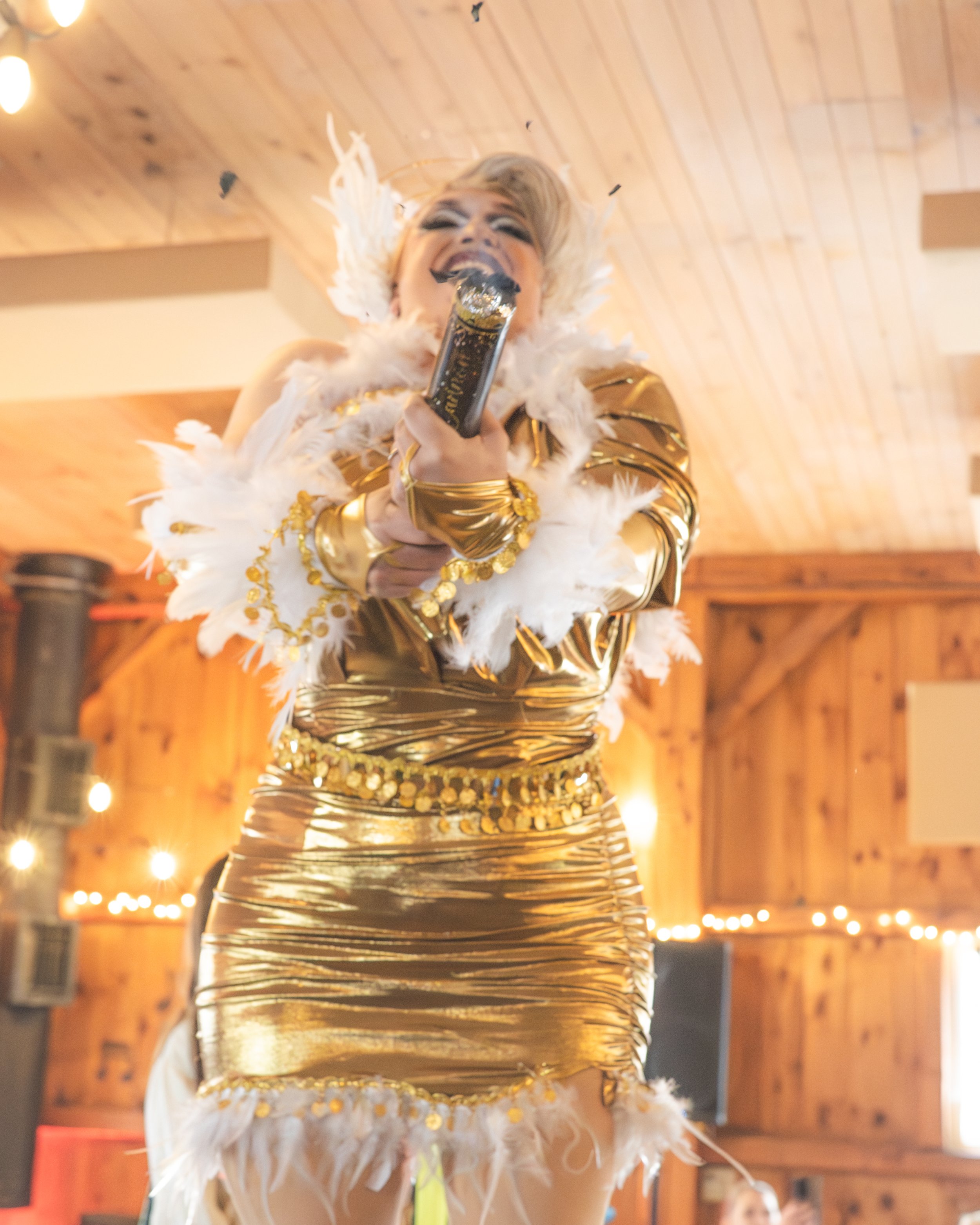 A woman in a shiny gold dress decorated with sequins and white feathers is singing into a microphone in a wooden-paneled room with string lights.