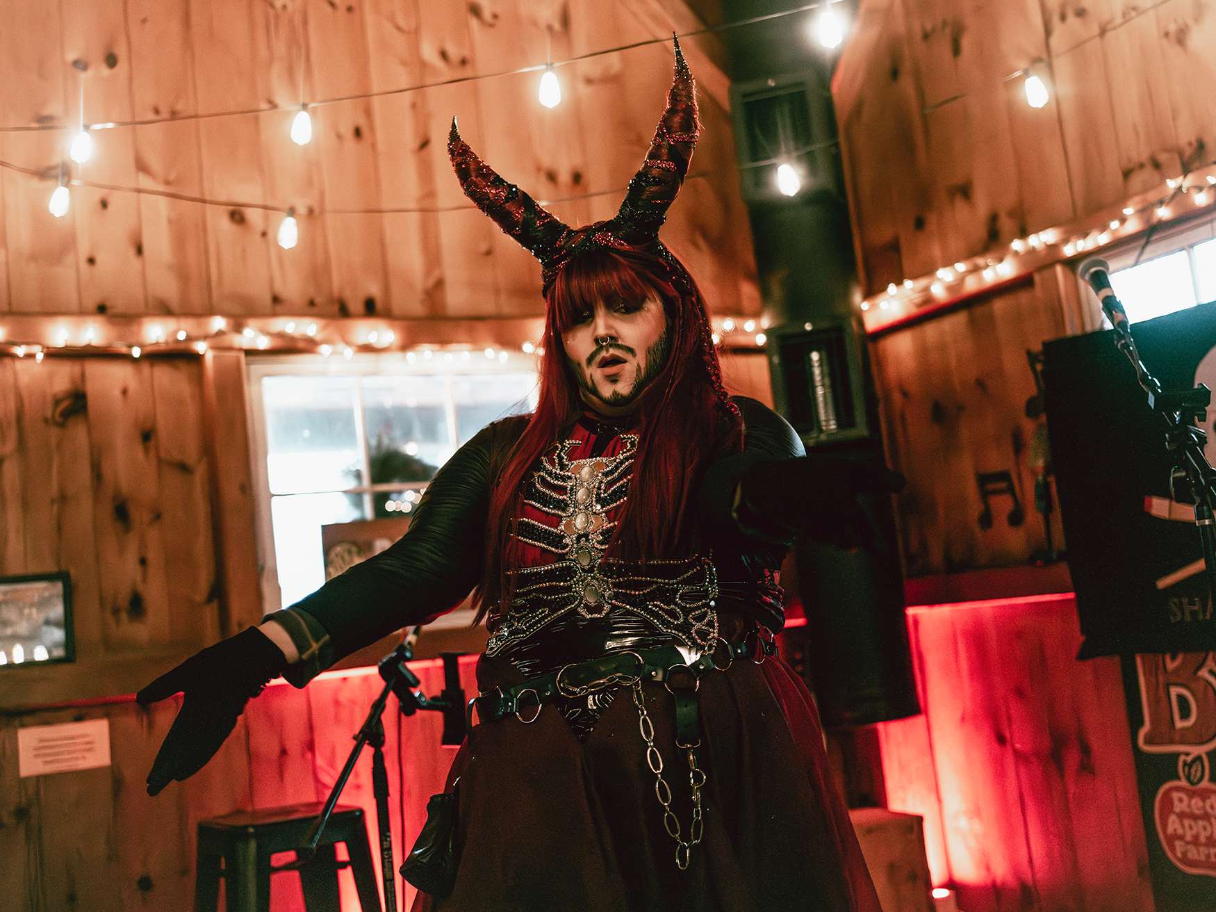 Person dressed in gothic costume with devil horns, long red hair, and skeleton jewelry, performing or posing in a rustic bar or music venue with string lights decorating the wooden walls.