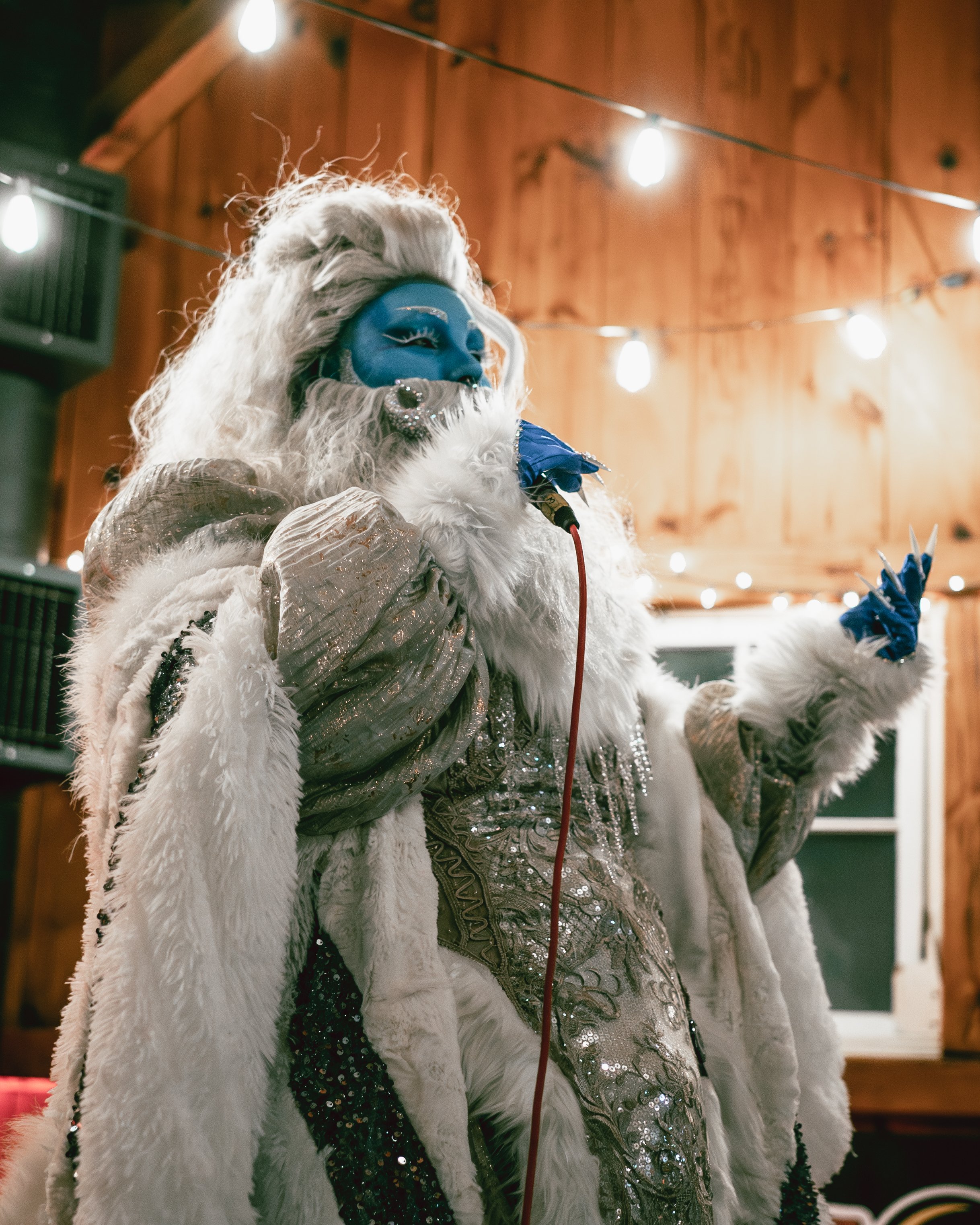 A person dressed as a winter holiday character, wearing a luxurious fur cape, sequined dress, and blue face paint, holding a microphone and singing in a warmly lit indoor setting with wooden walls and string lights overhead.