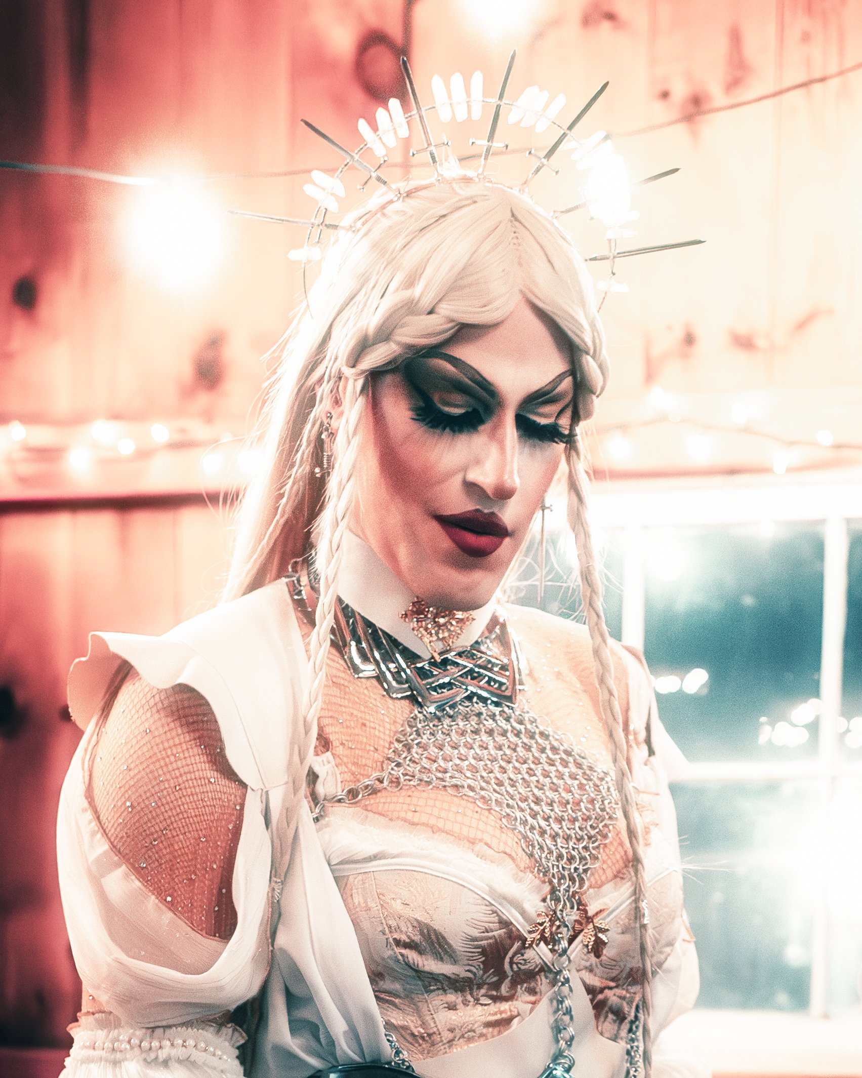 A woman with platinum blonde hair styled in braids, wearing dark makeup, jewelry, and a detailed outfit, standing in a warmly lit room with wooden walls and string lights.