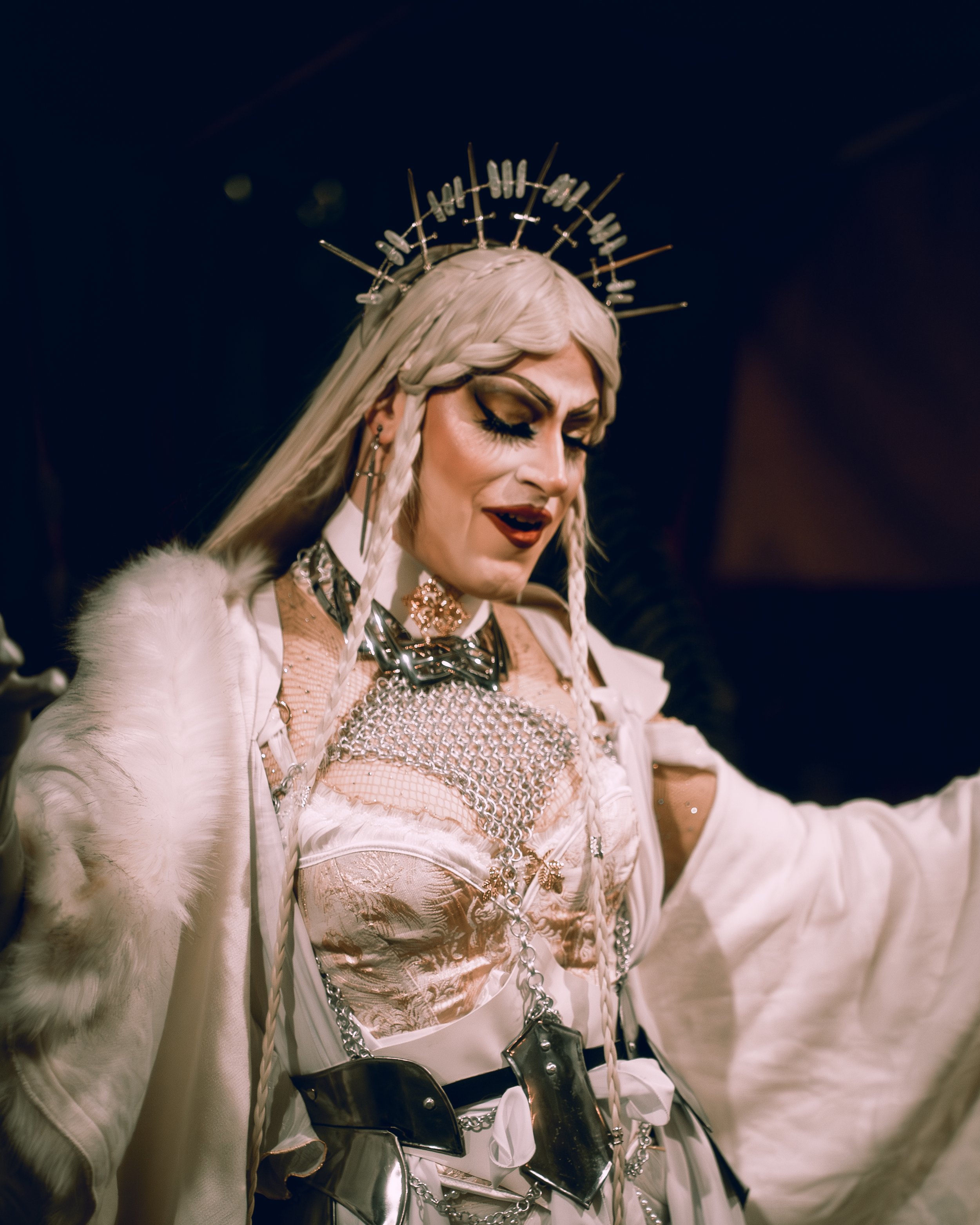 A woman dressed in elaborate, fantasy-inspired costume with metallic accessories, a crown-like headpiece, and long braided platinum blonde hair.
