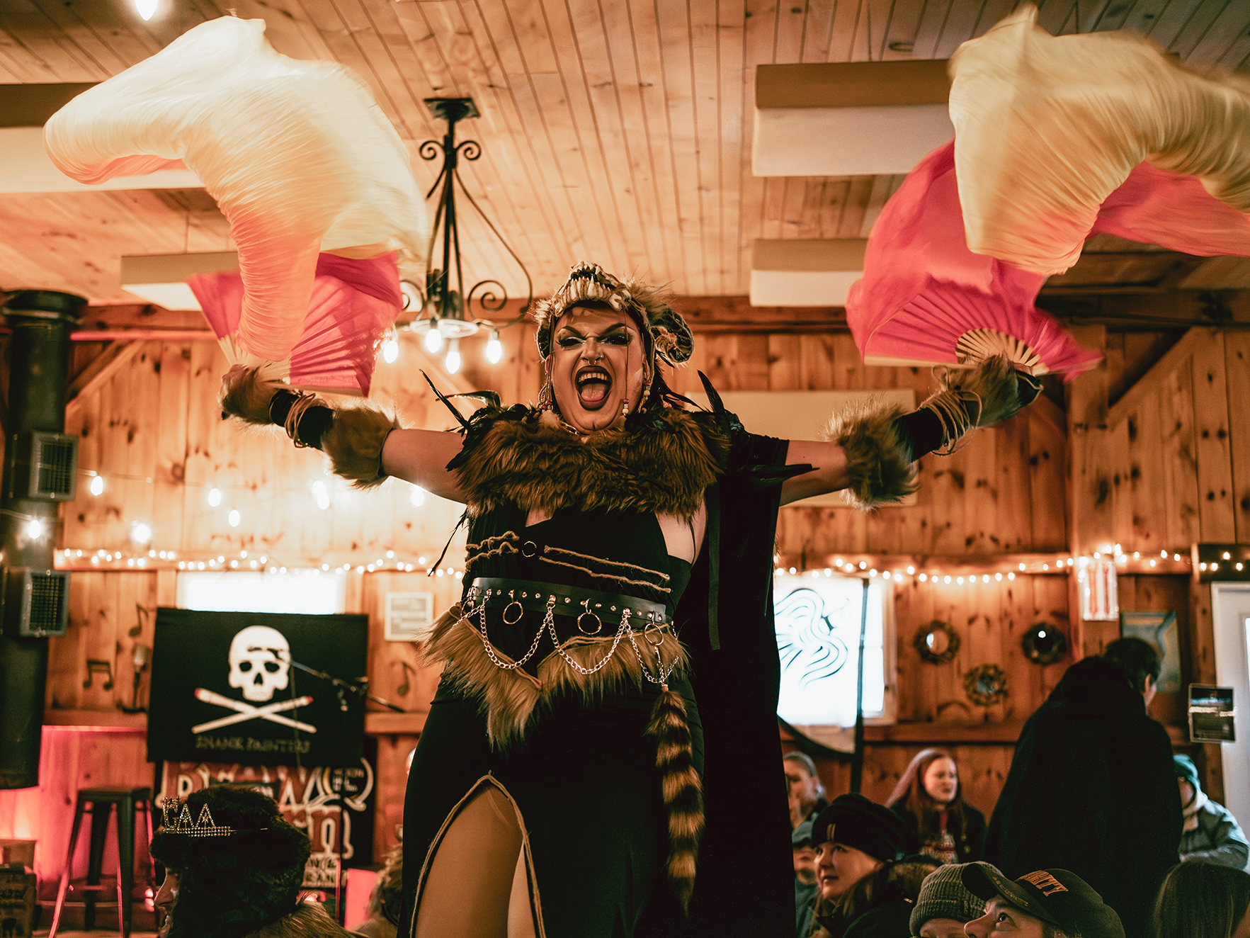 Performer in a costume with animal fur and chains, holding large pink and white fans, singing or shouting with a wide open mouth at a lively indoor event with wood-paneled walls and string lights.