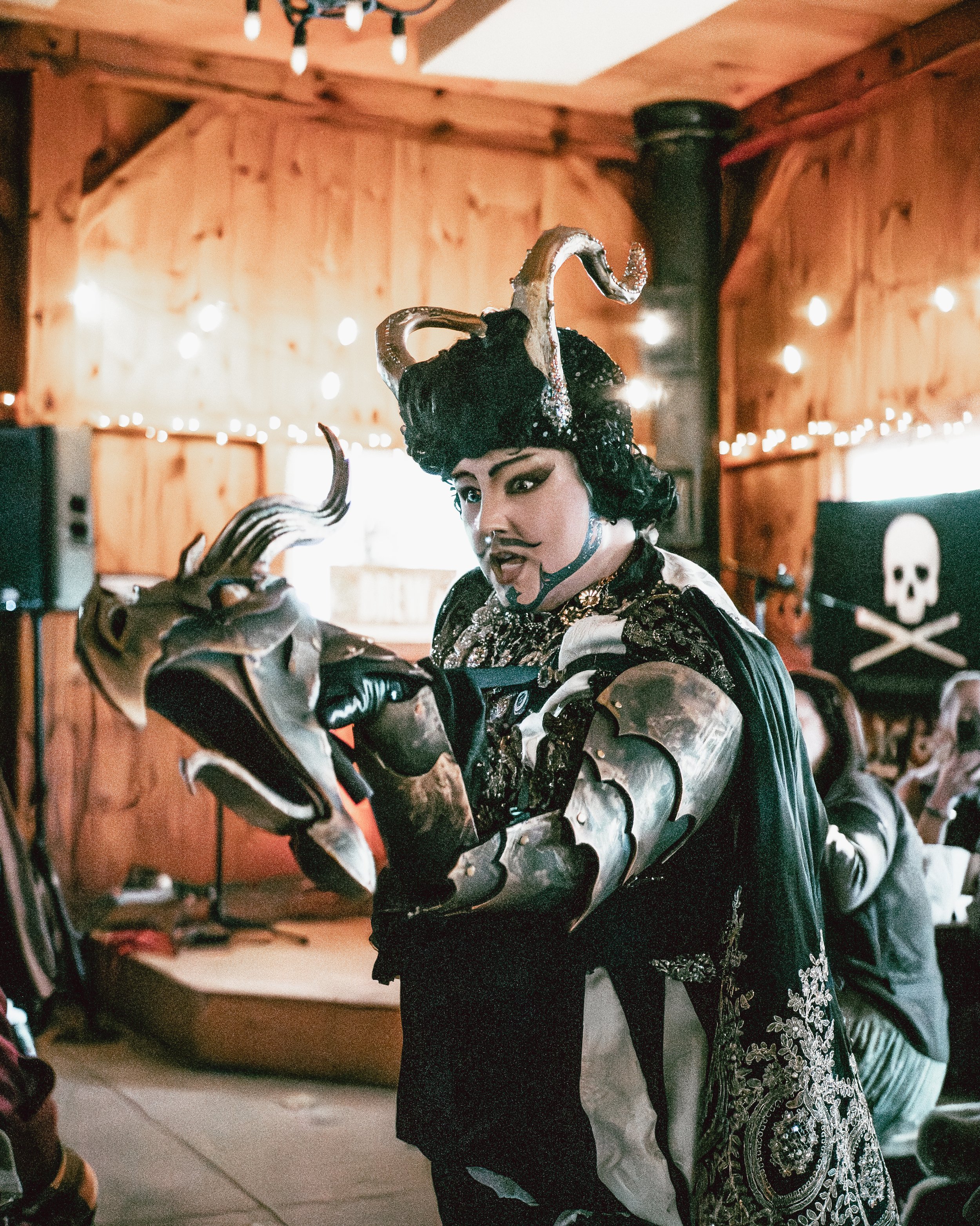 Person dressed as a fantasy villain with metallic armor, ornate black clothing, and a headpiece with tentacle-like adornments, holding a decorative sword in a wooden room with string lights and a pirate flag.