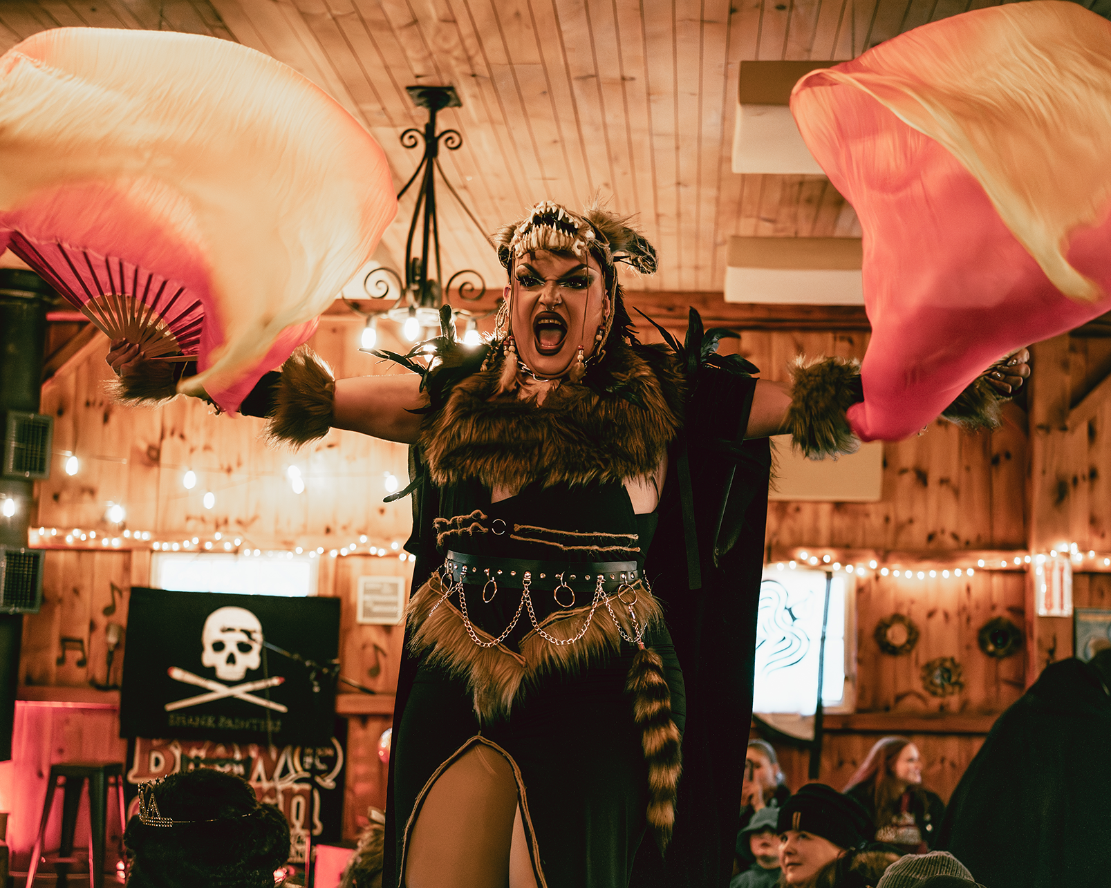 A performer dressed as a fierce tiger queen with animal print and chains, holding pink and orange fans, mid-performance in a wooden hall decorated with string lights and a pirate-themed backdrop.
