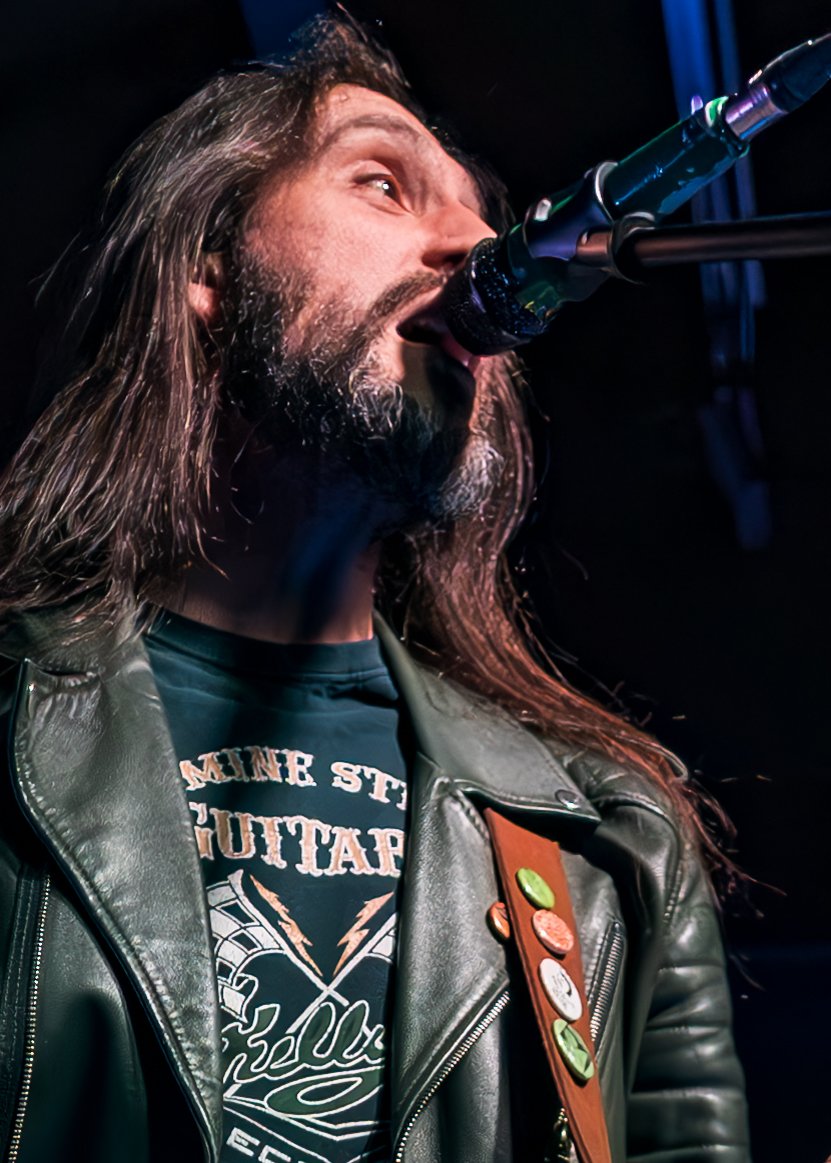 Male musician with long hair and beard singing into a microphone on stage, wearing a leather jacket with colorful pins and a graphic T-shirt.