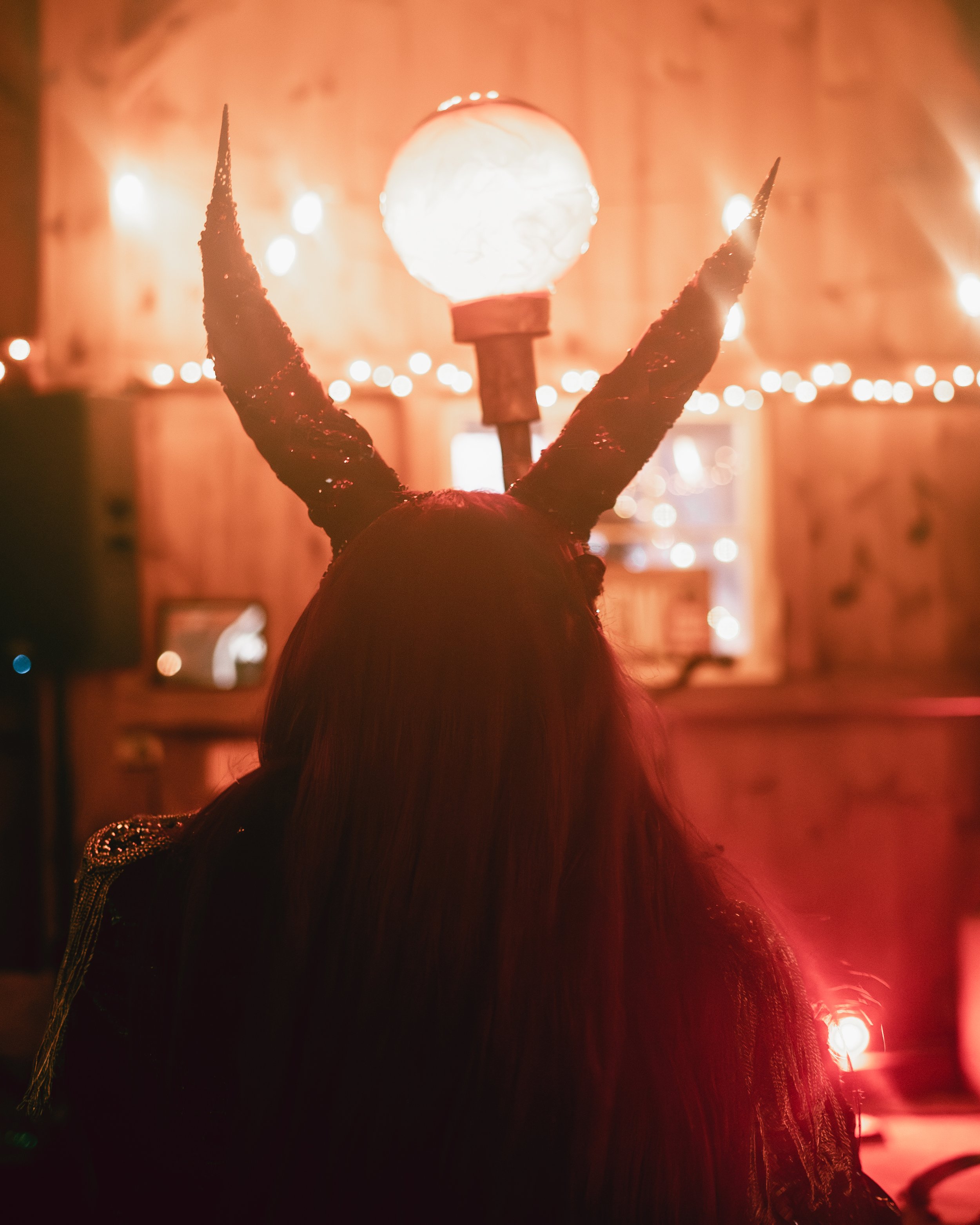 Person with devil horns headband and a moon lamp in the background at a dimly lit indoor event.