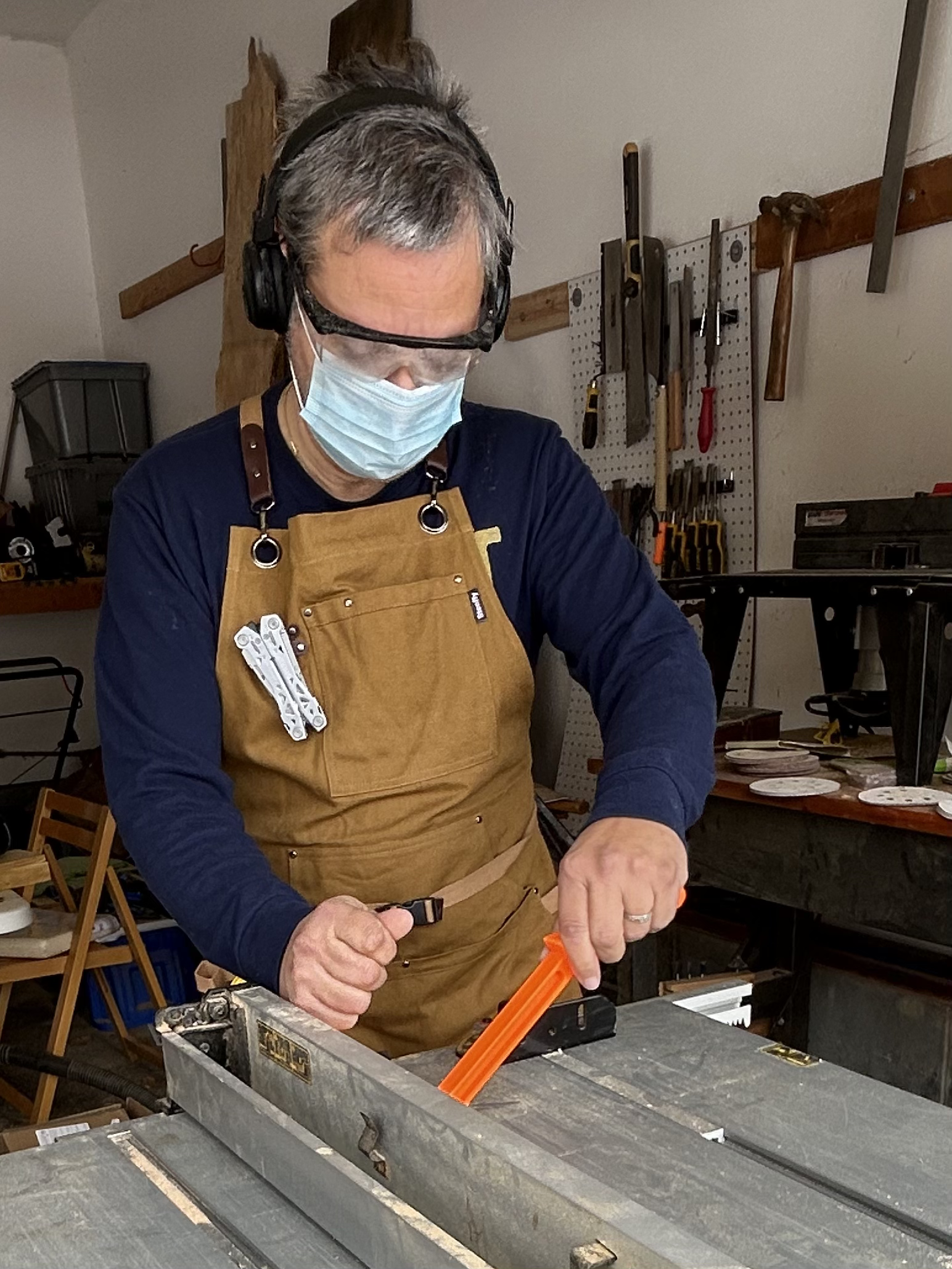 An older man wearing safety goggles, a face mask, and a brown apron is working in a woodworking workshop. He is using a handsaw on a piece of wood.