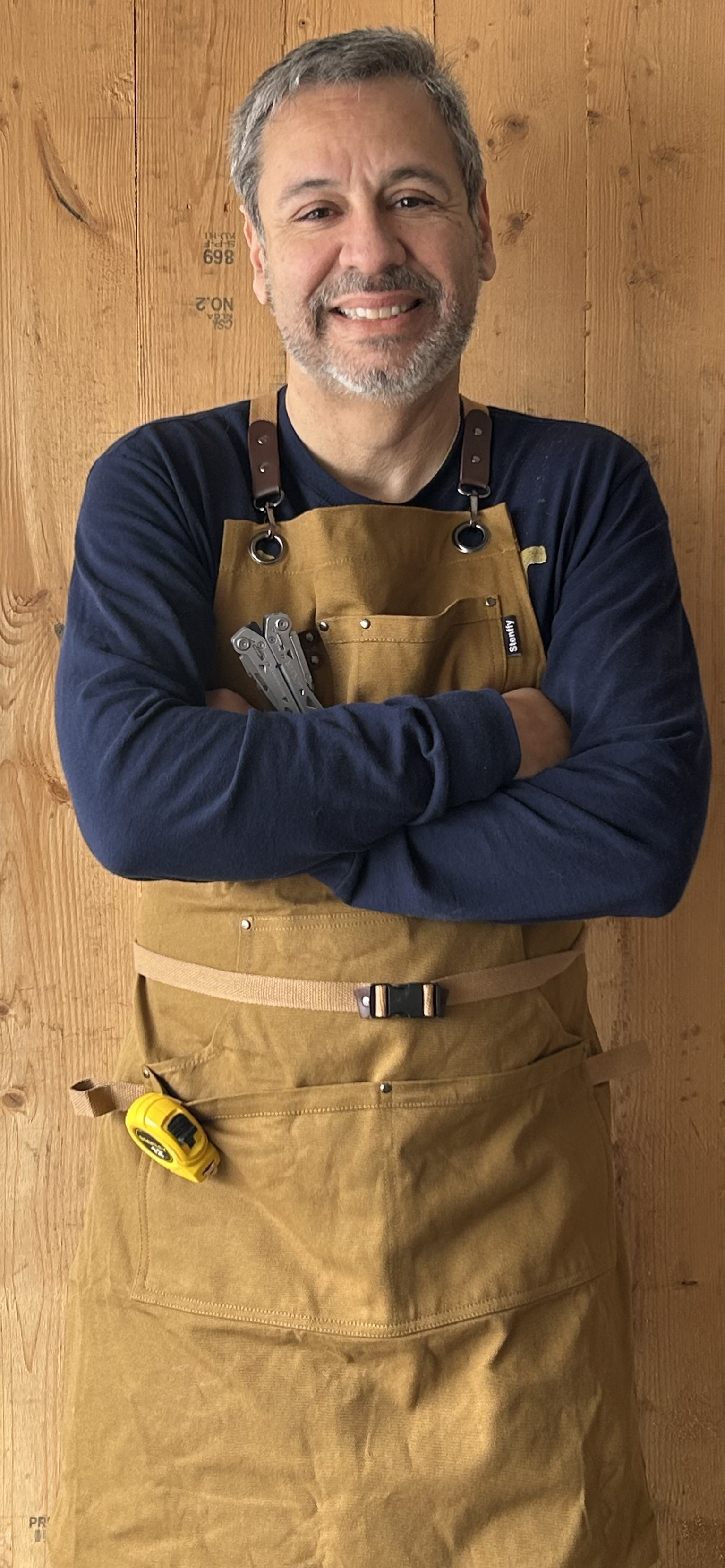 A man with gray hair and beard smiling, standing in front of a wooden wall, wearing a blue long sleeve shirt and a brown apron with a tape measure attached, holding a multi-tool.
