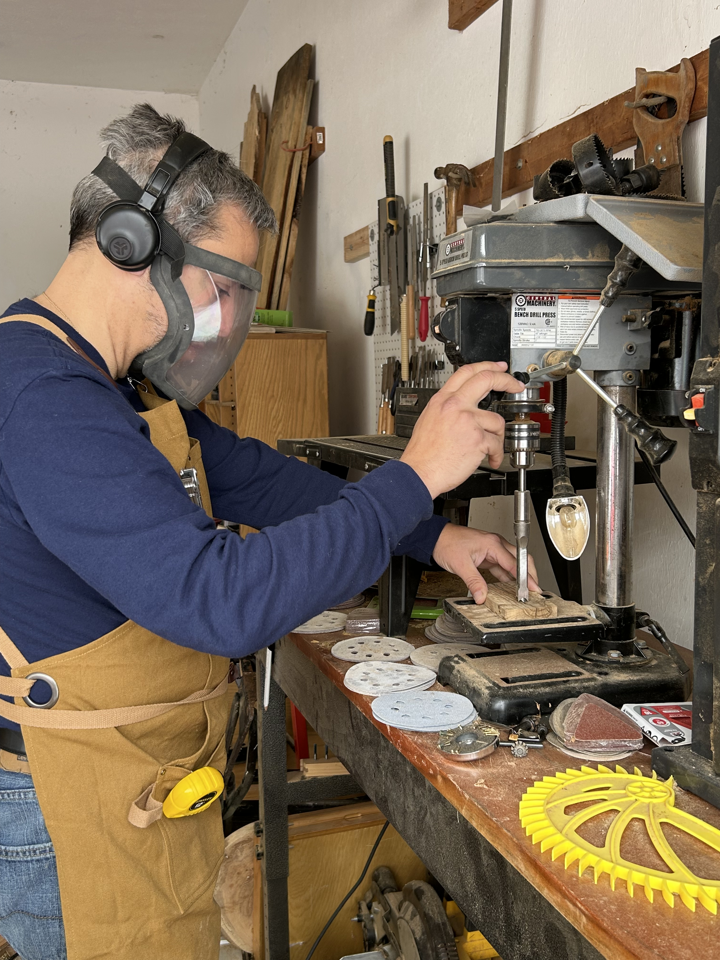 A man wearing a face shield, ear protection, and a brown apron is operating a drill press in a woodworking workshop.