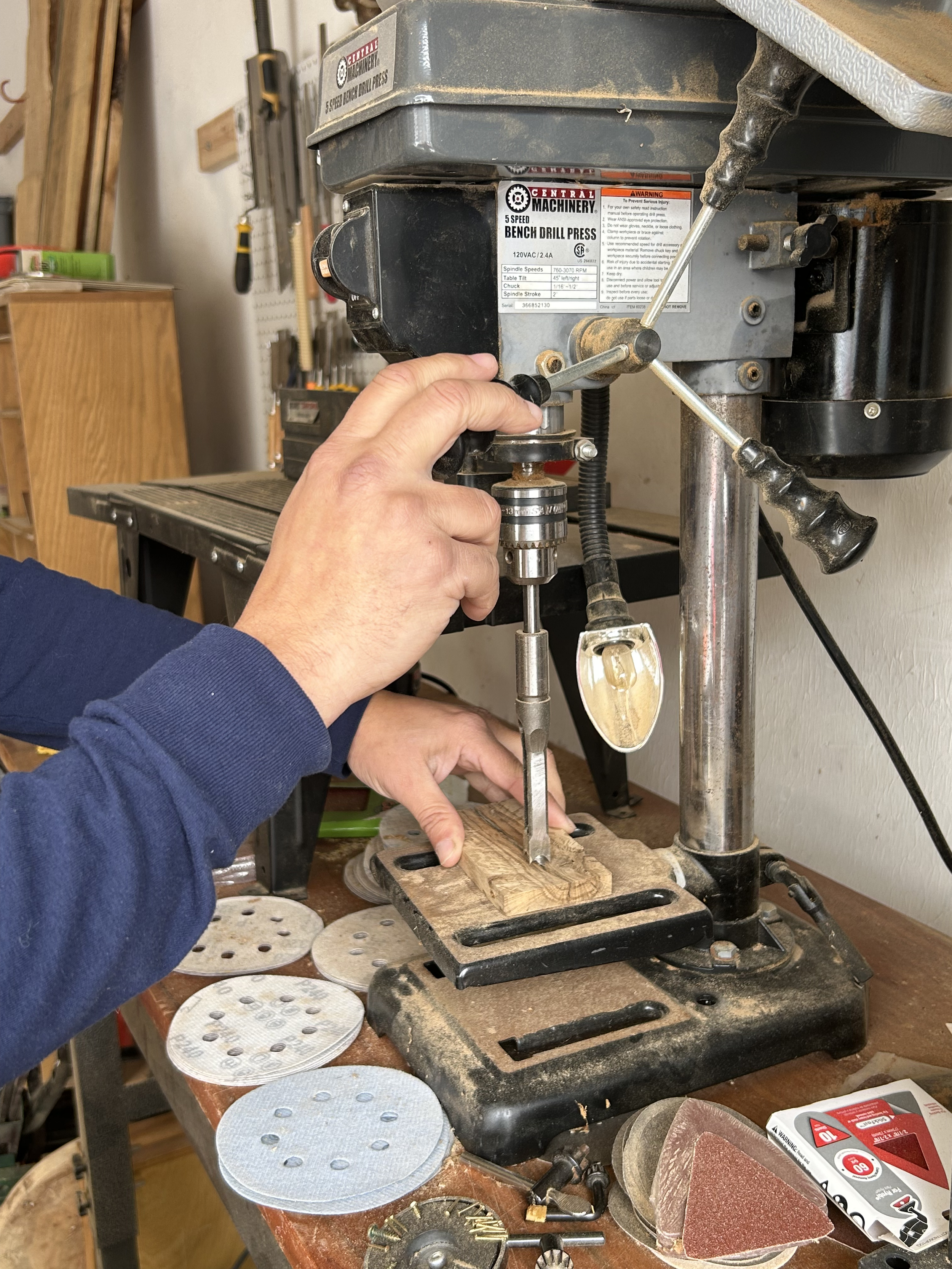 A person using a benchtop drill press to drill into a piece of wood in a workshop.