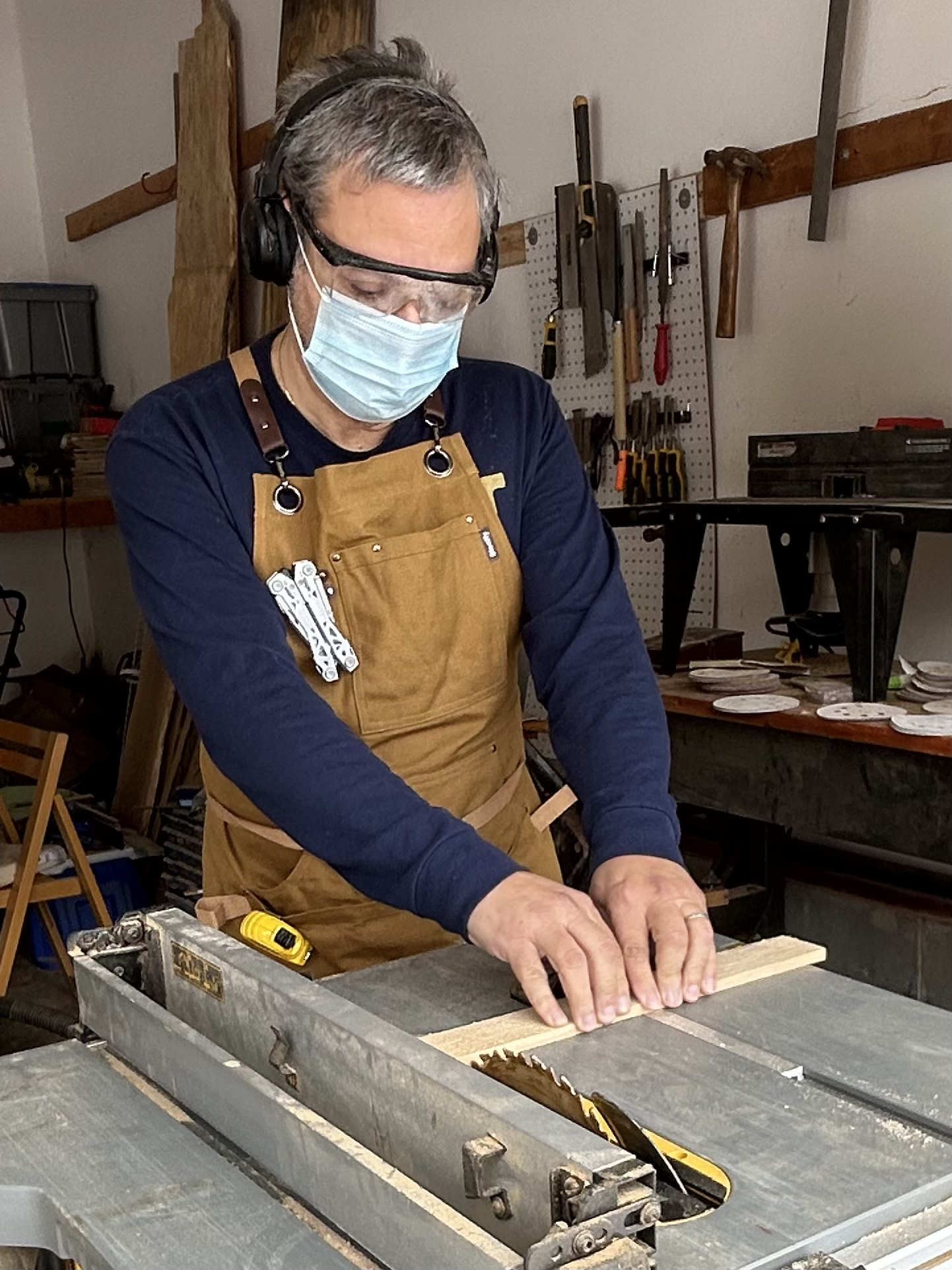 A man working with a table saw in a woodworking shop, wearing safety goggles, a face mask, headphones, and an apron, cutting a piece of wood.