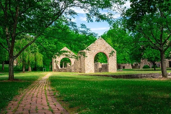 A red‑brick pathway leads through bright green grass toward the stone ruins of the historic carpenter’s shop at Lock Ridge Park, where weathered fieldstone walls with tall arched openings stand against a backdrop of leafy summer trees and blue sky.