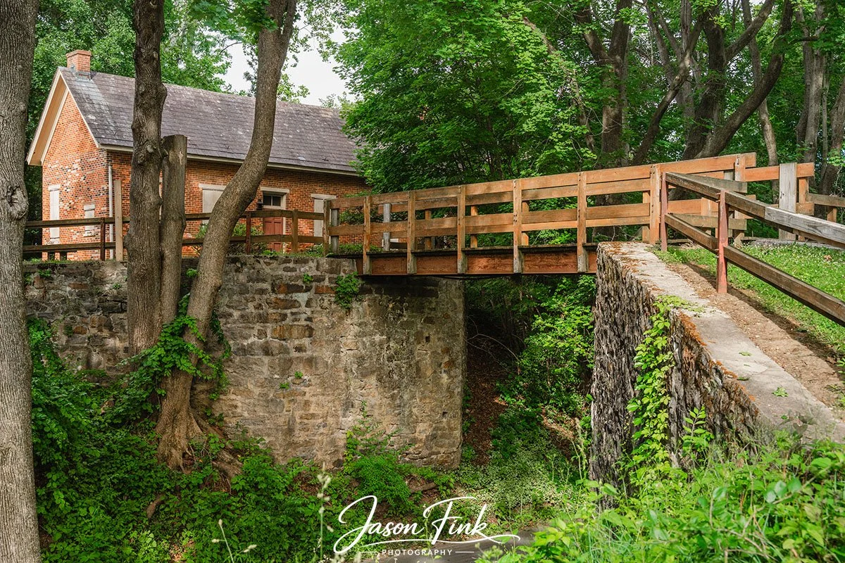 Soft light settles over the wooden bridge and the old weighmasters house at Lock Ridge Park, giving the scene a quiet, almost storybook calm. The weathered stonework and hand-built textures speak to the parks industrial past, when this property forme