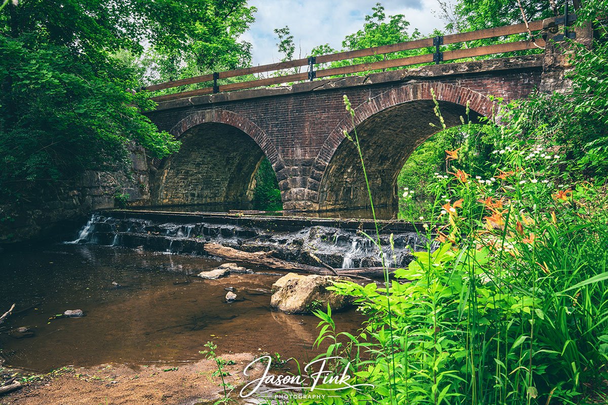Swabia Creek winds softly beneath the old stone arches, carrying the quiet history of Lock Ridge Park in its gentle flow. The bridge rises with warm, weathered brick, its wooden rail catching the afternoon light as water tumbles over rock ledges belo