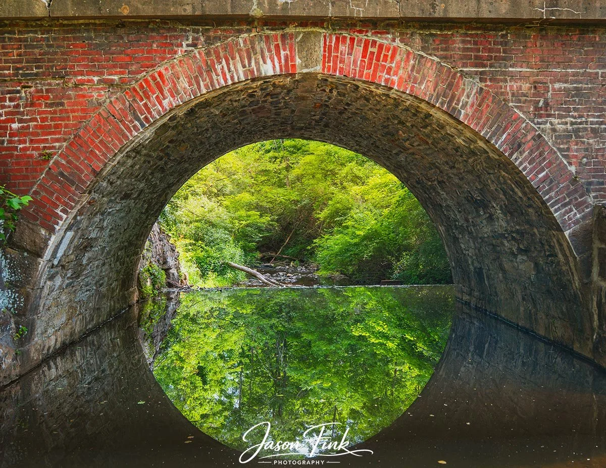 A redbrick arch rises over still waters of the Swabia Creek in this quiet corner of Lock Ridge Park, its reflection forming a near perfect mirror beneath the curve. The mix of brick and dark stone hints at the site's industrial origins, when the Lock
