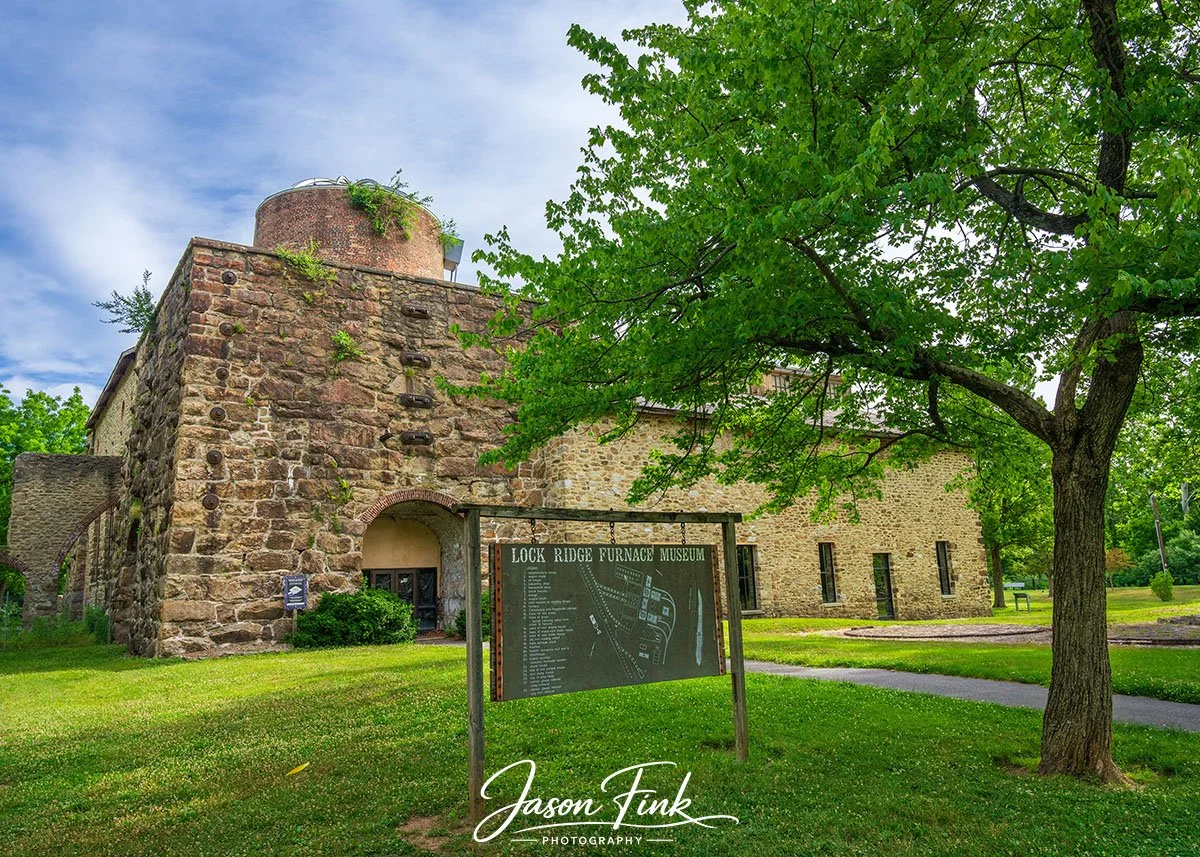 The Lock Ridge Furnace Museum stands here like a stone bound memory of Alburtis industrial past, its massive masonry walls and towering stack rising from the green landscape with a quiet, enduring presence. The weathered sign in the foreground feels 