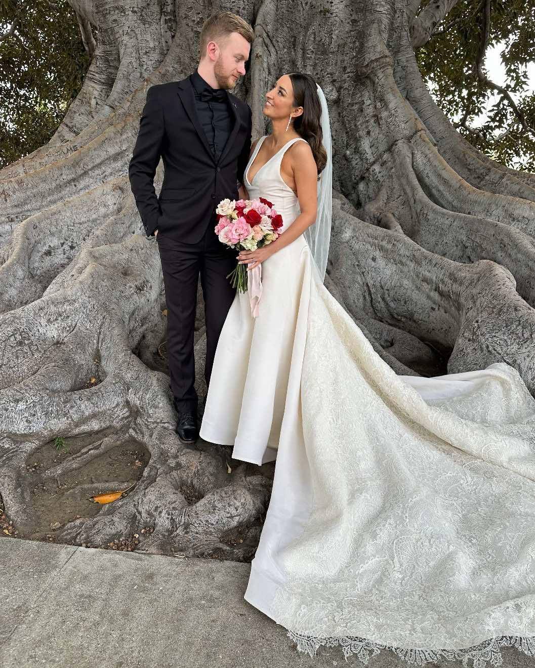 A bride and groom standing in front of a large tree, gazing at each other. The bride is holding a bouquet of pink, red, and white flowers, wearing a white gown with a long train and veil. The groom is in a black tuxedo with a black shirt, standing wi