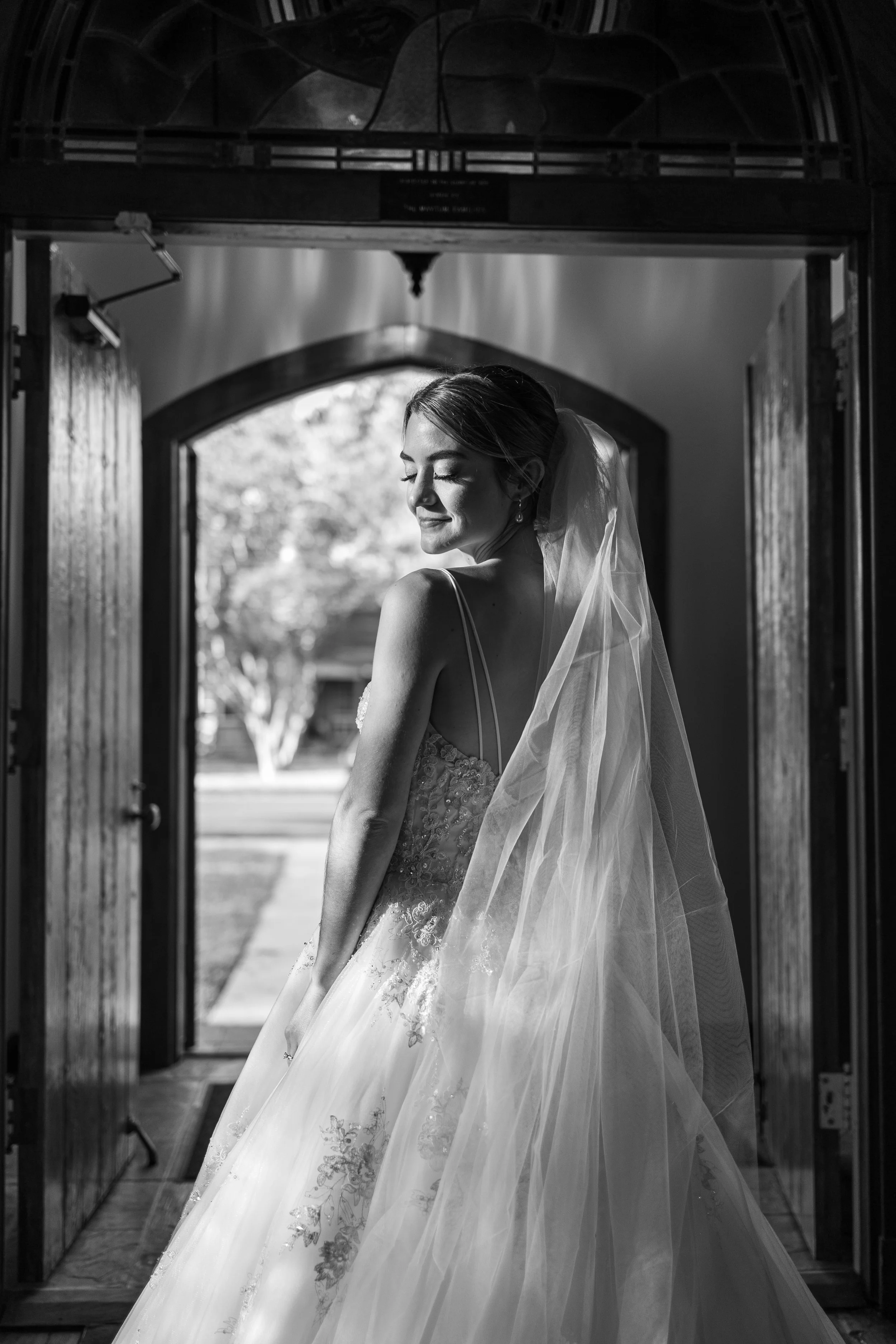 A bride in a wedding gown and veil standing in a doorway, smiling with eyes closed in black and white.