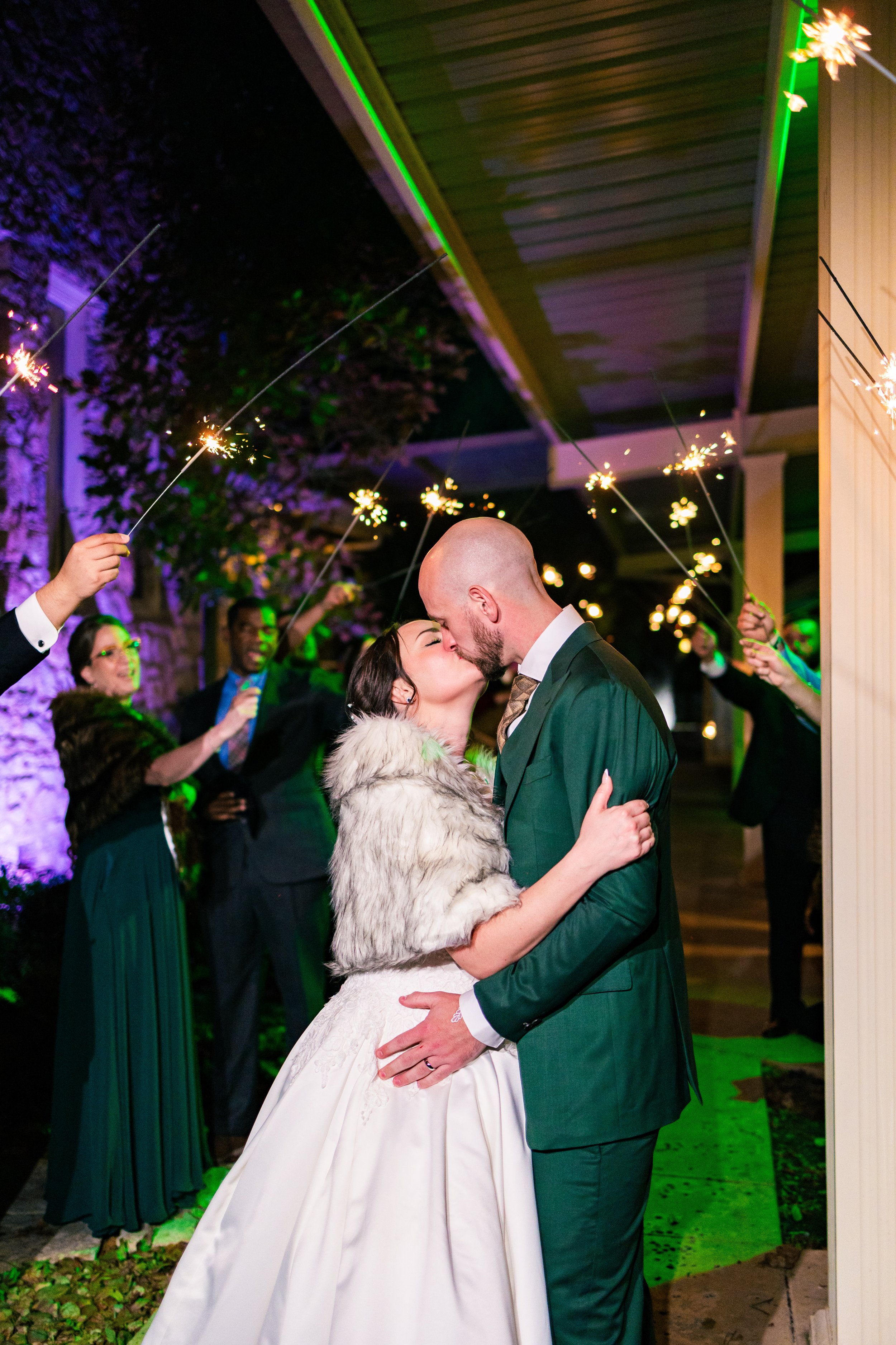 A newlywed couple sharing a kiss at their wedding celebration, surrounded by guests holding sparklers, with green and purple lighting.