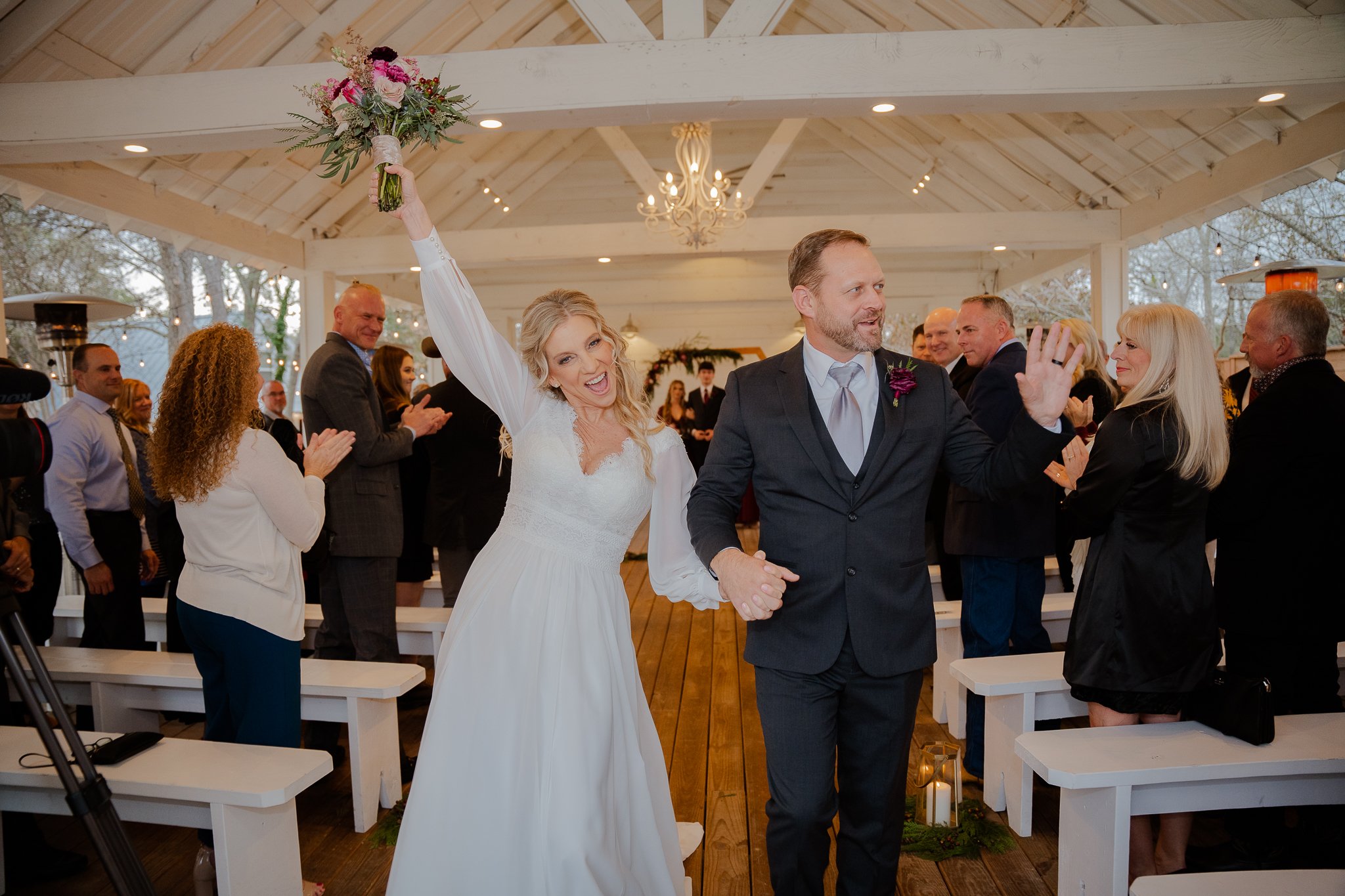 Bride and groom celebrating after wedding ceremony, holding hands and smiling, with guests clapping and cheering in a decorated outdoor pavilion.