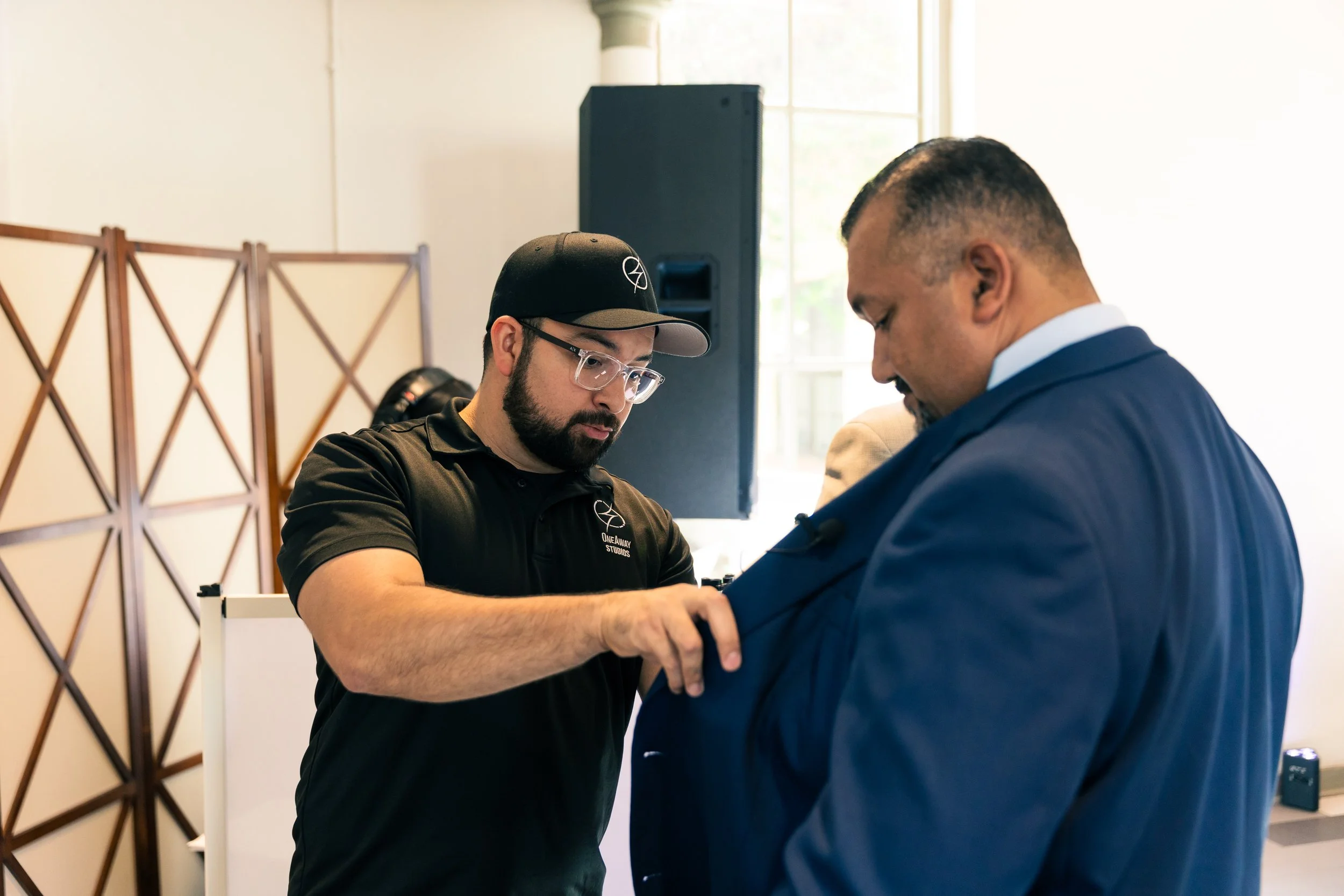 A man in a black polo shirt and cap helps a man in a suit put on his jacket in an indoor setting with large windows and wooden room divider.