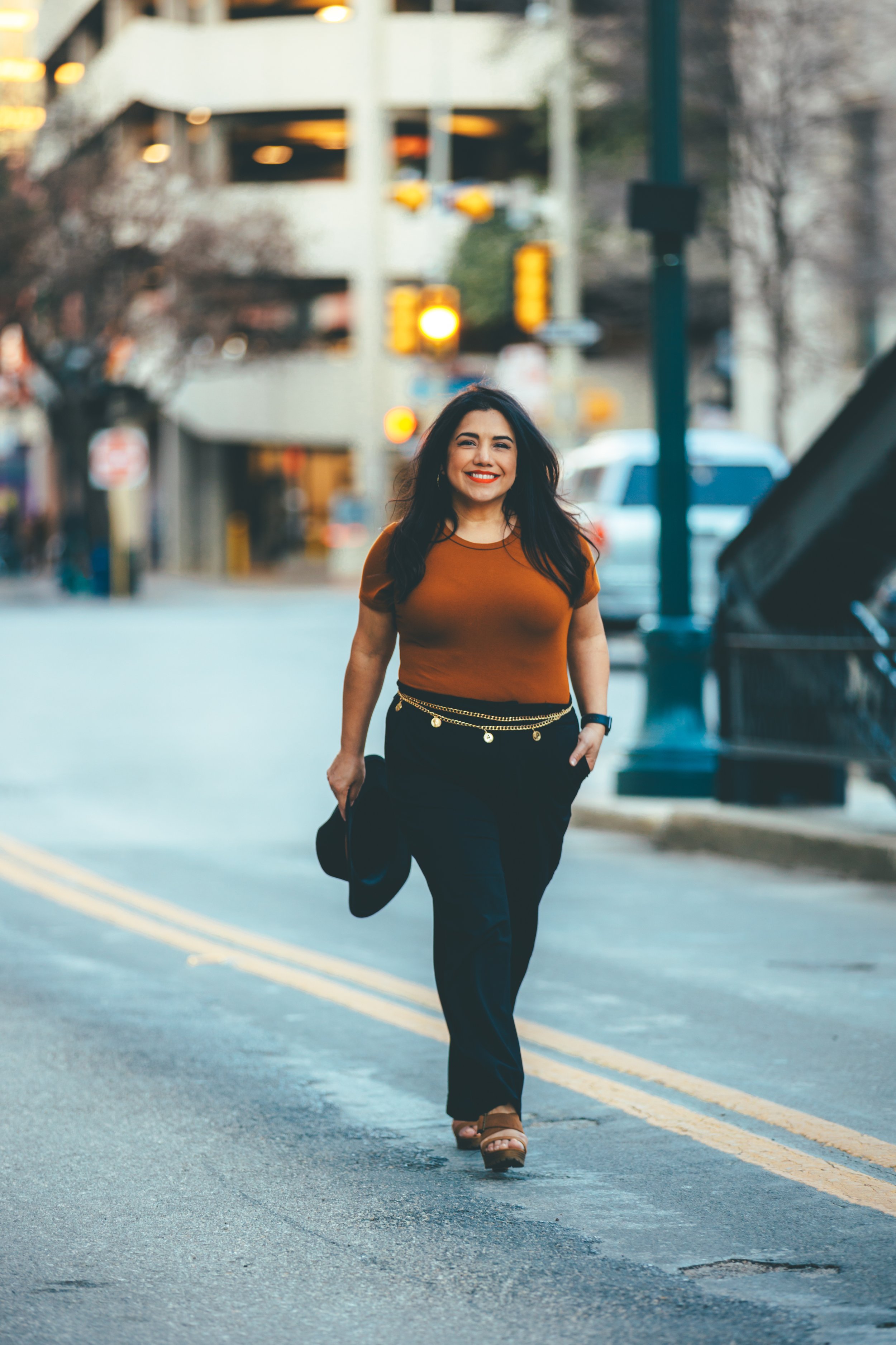 Woman walking on city street smiling, holding a black hat, dressed in a brown top and black pants.