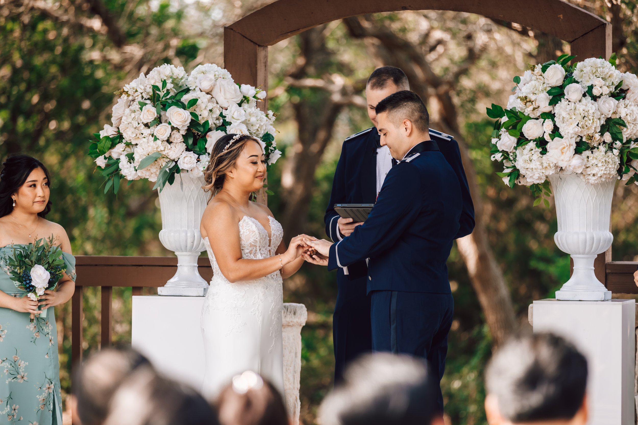 A couple getting married in an outdoor ceremony, exchanging rings under a wooden arch decorated with large white flower arrangements, with an officiant and a bridesmaid holding a bouquet nearby.