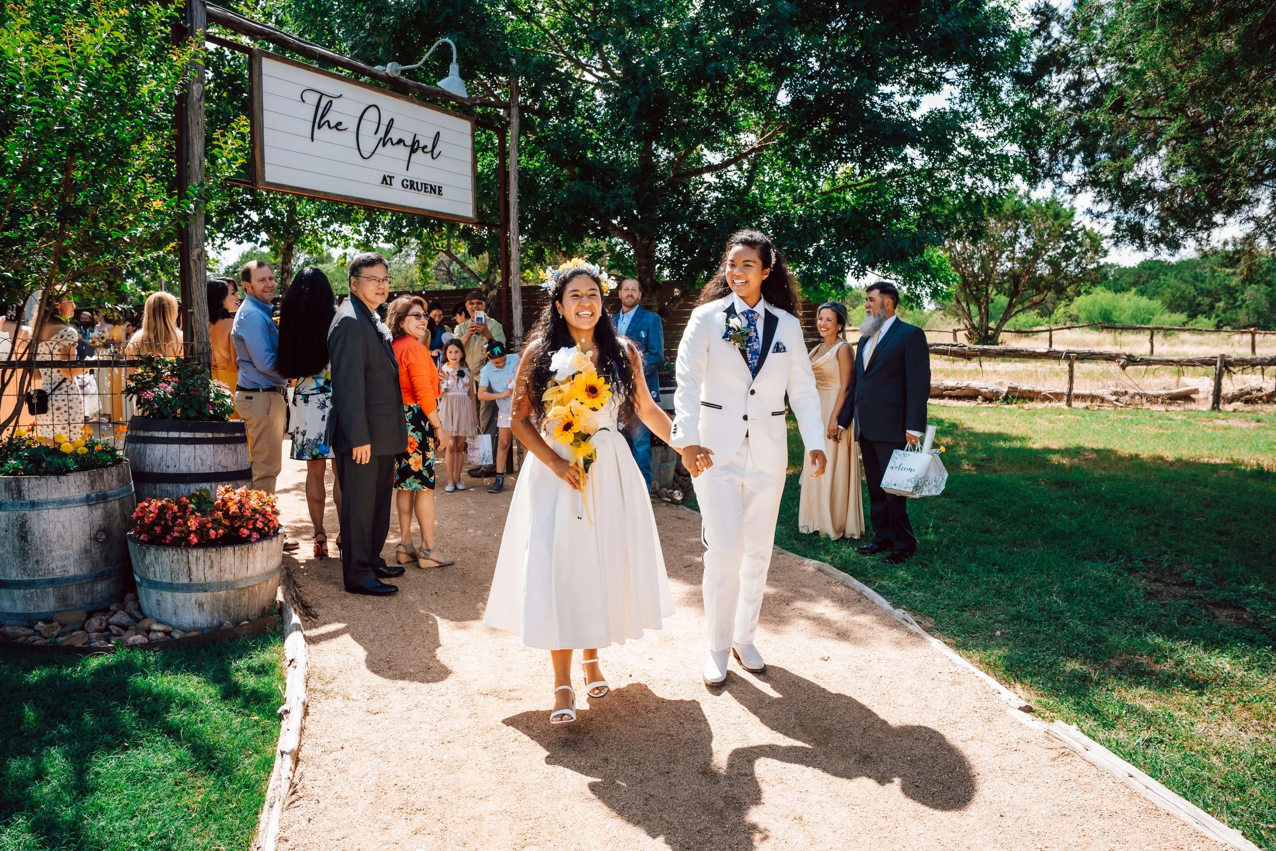 A same-sex couple, dressed in wedding attire, walking hand-in-hand outdoors during a wedding ceremony, with guests in the background under a large tree.