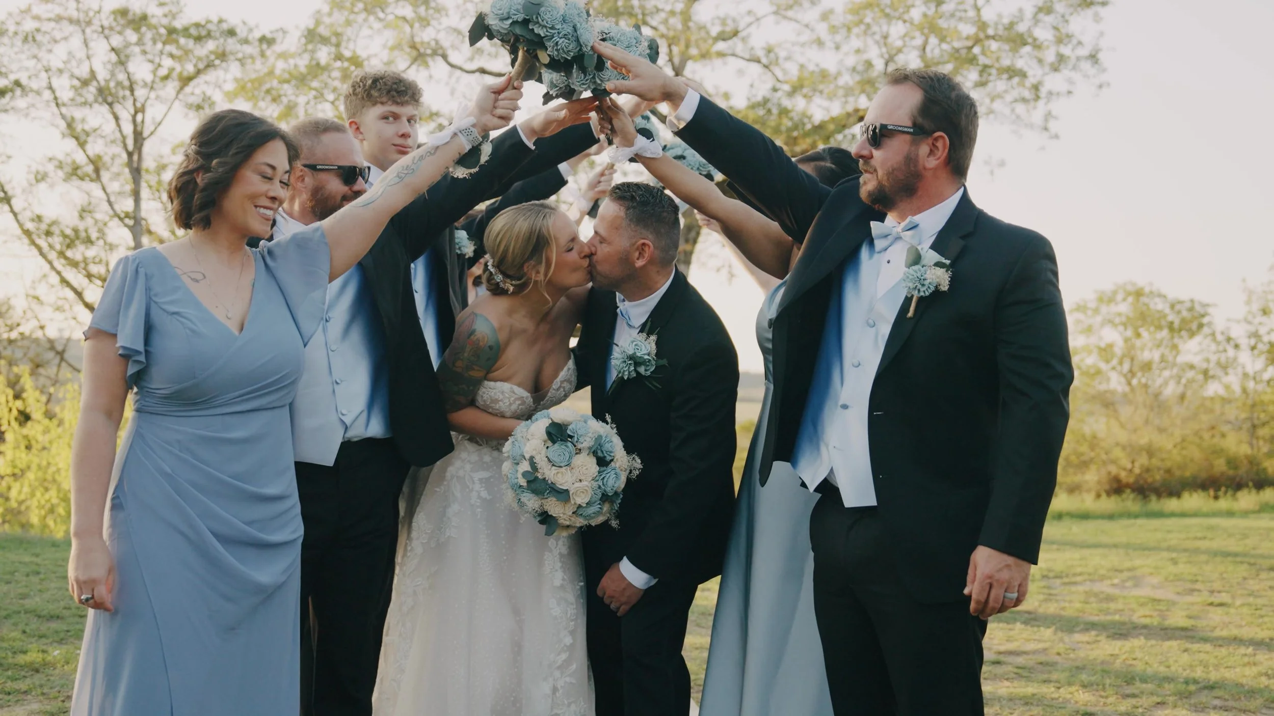 A newly married couple shares a kiss while surrounded by friends and family holding a floral arrangement overhead at an outdoor wedding ceremony in the late afternoon.