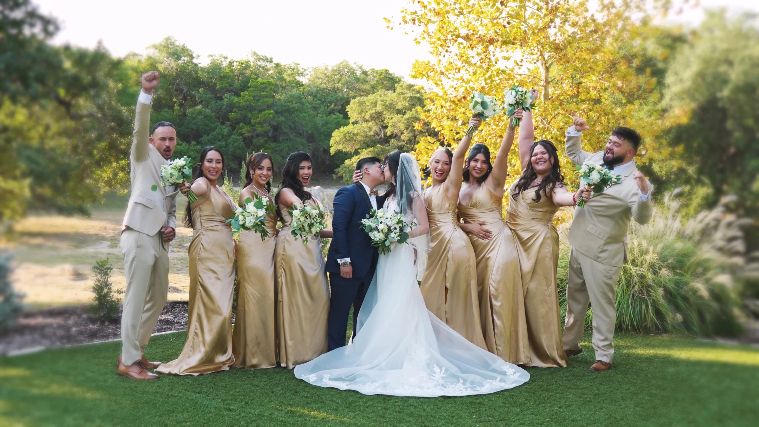 A wedding party outdoors with the bride and groom kissing in the center, surrounded by bridesmaids and groomsmen celebrating with bouquets and raised arms, in a lush green setting with trees and sunlight.