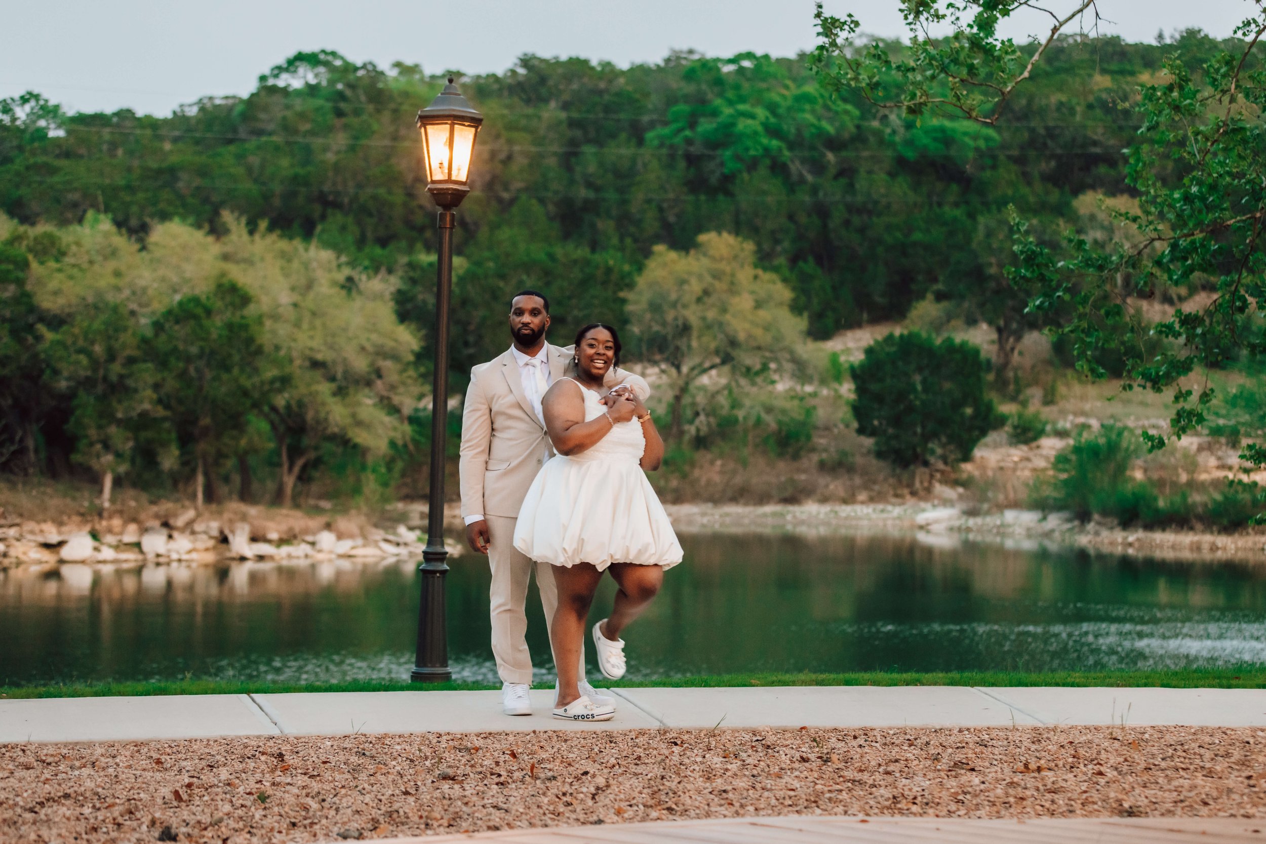 A happy couple in formal attire posing outdoors near a body of water during the evening, with trees and hills in the background and a lamp post nearby.