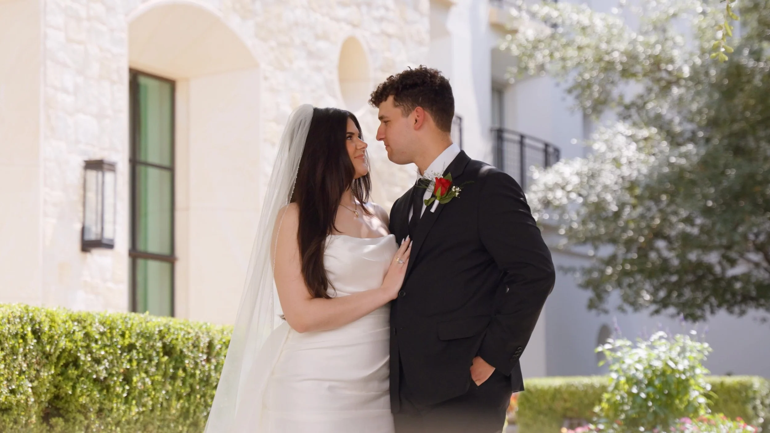 A newlywed couple standing close together outdoors, gazing into each other's eyes, with a stone building and greenery in the background.