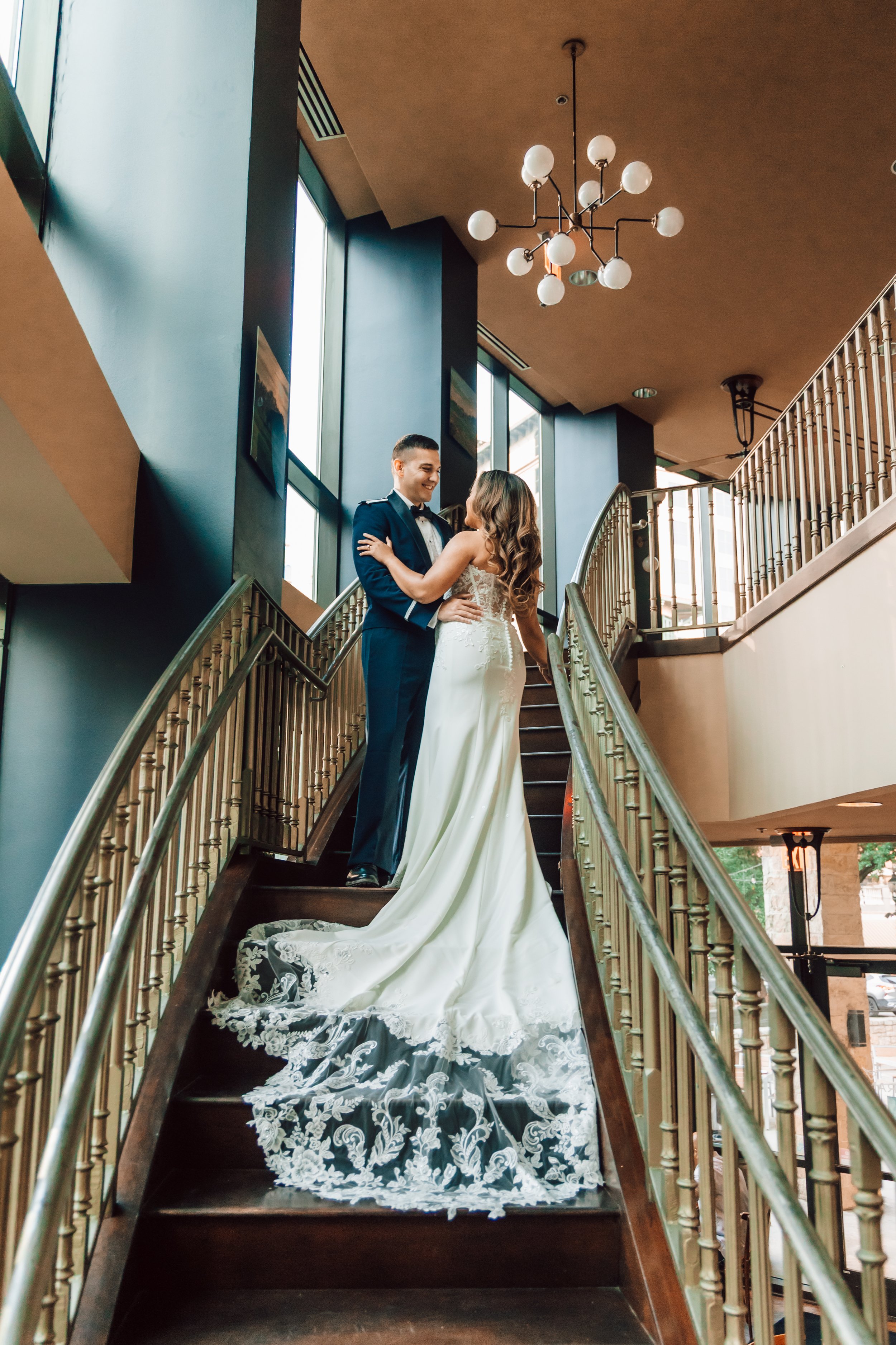 A bride and groom dancing on a staircase inside a modern building with large windows and hanging lights.