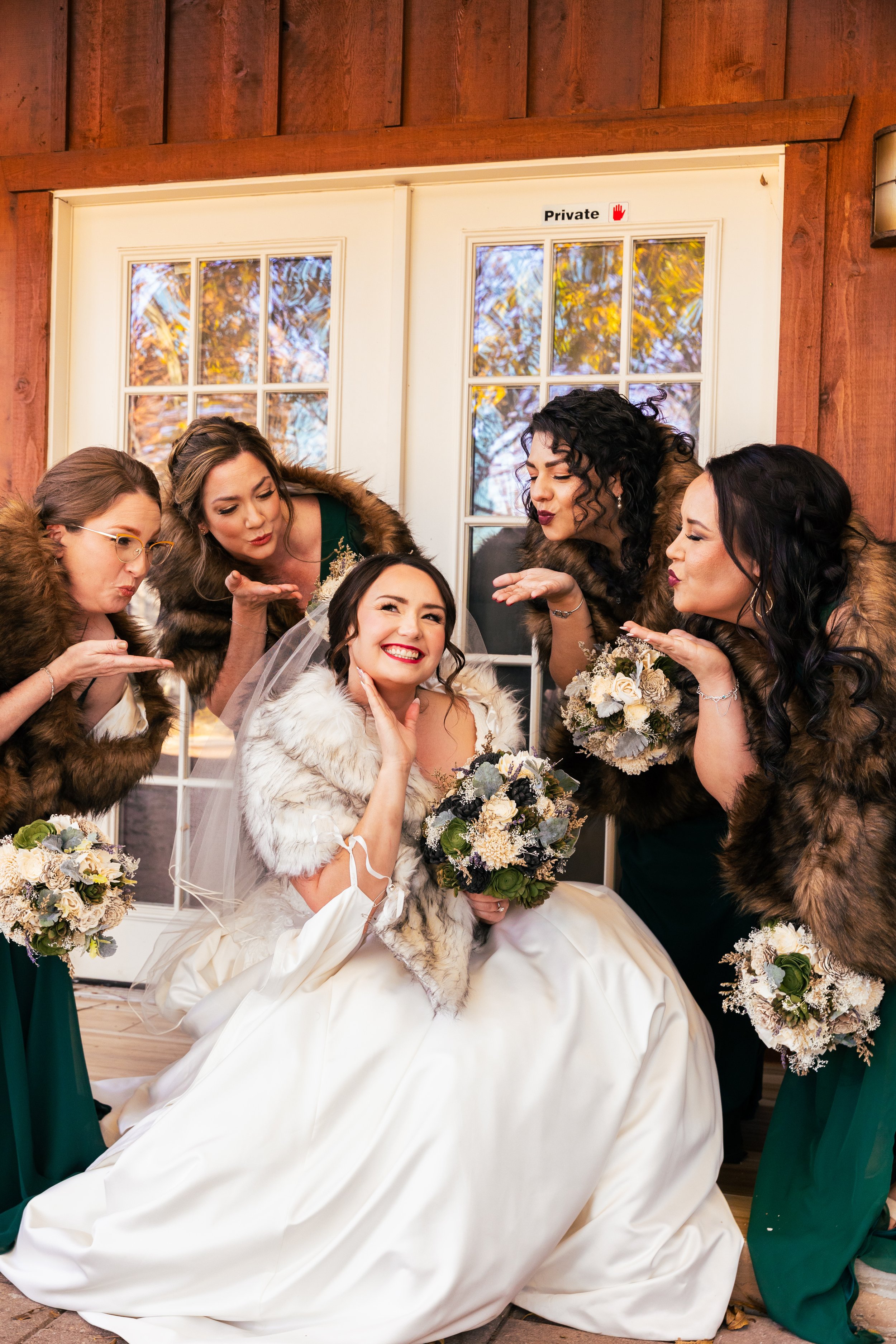 Bride seated in wedding dress and white fur coat, surrounded by four bridesmaids wearing green dresses and fur stoles, all holding bouquets, posing playfully for photos indoors with wooden walls.