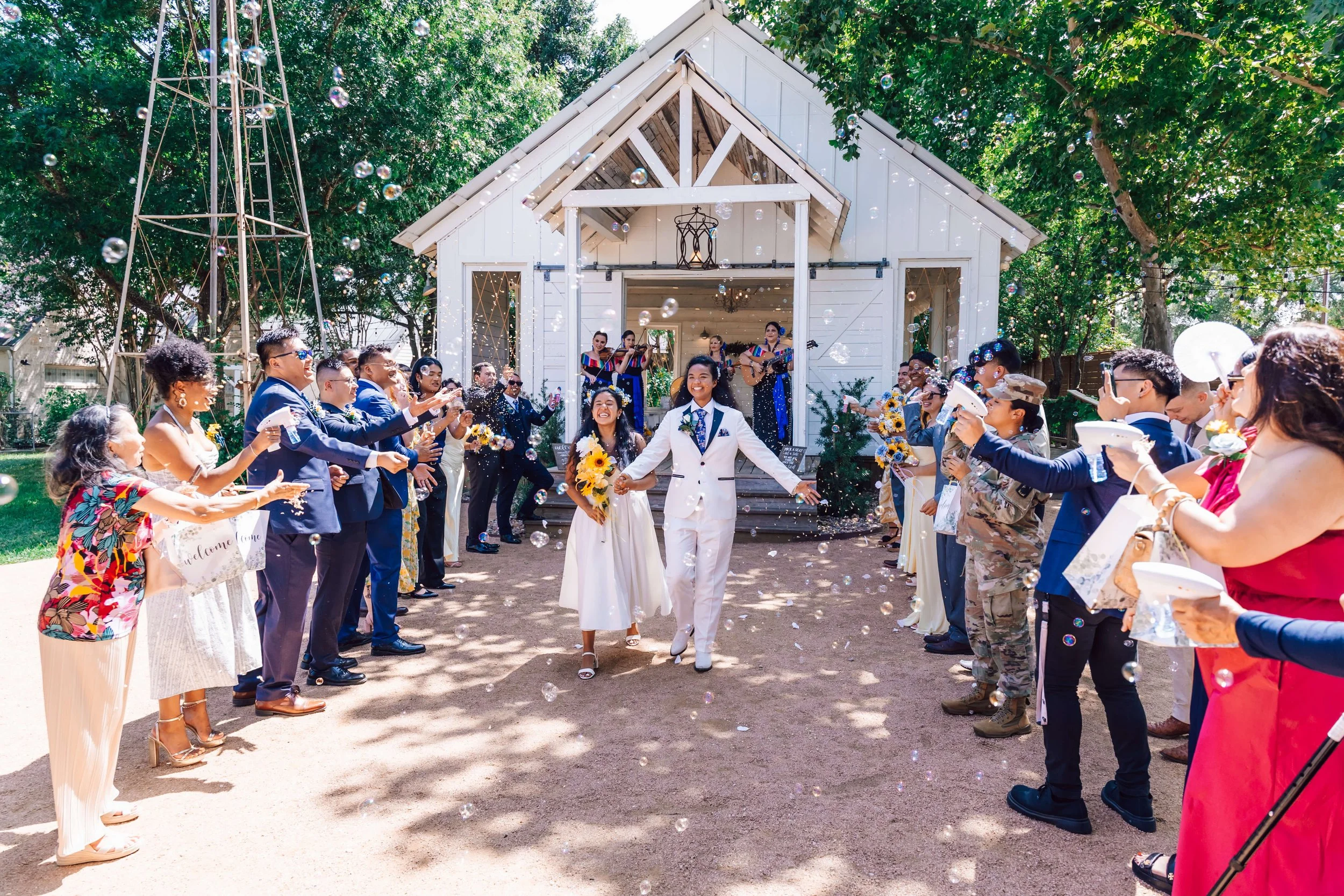Celebrating wedding of two women with guests throwing bubbles and confetti outside a white chapel.