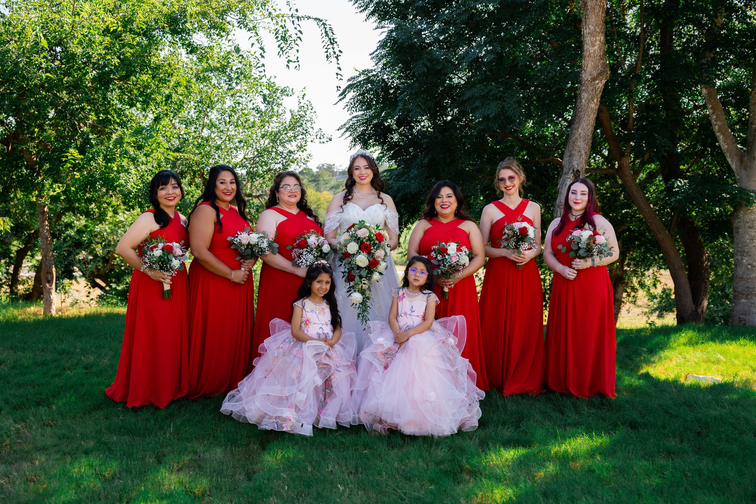 A bride and eight bridesmaids pose outdoors on green grass with trees in the background. The bride is in a white lace gown, holding a large bouquet, surrounded by women in red dresses holding smaller bouquets. Two young flower girls in pink and white