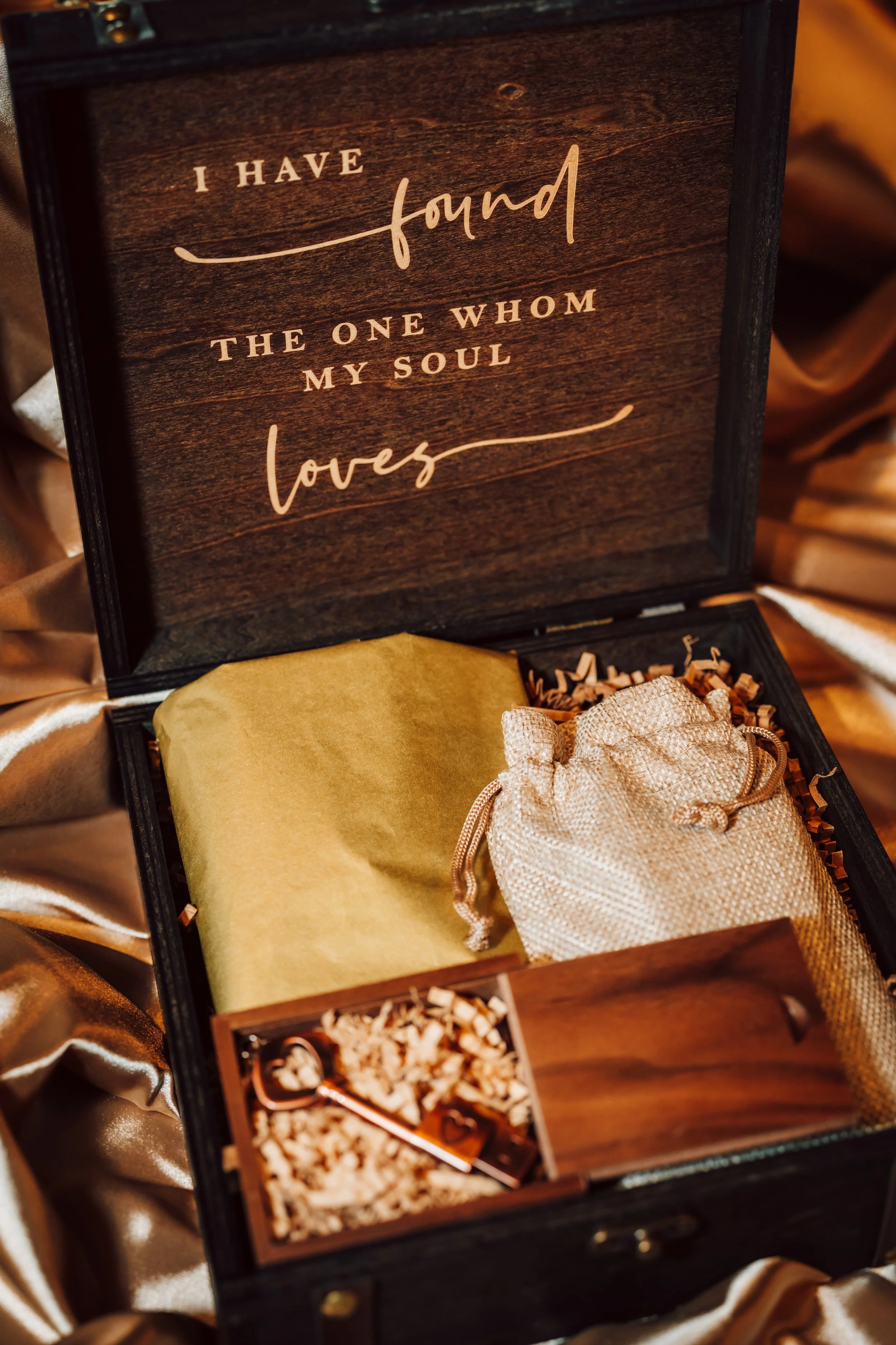 A gift box with a sentimental quote on the inside lid, containing a wrapped gift, a small pouch, and a decorative key, with gold satin fabric in the background.